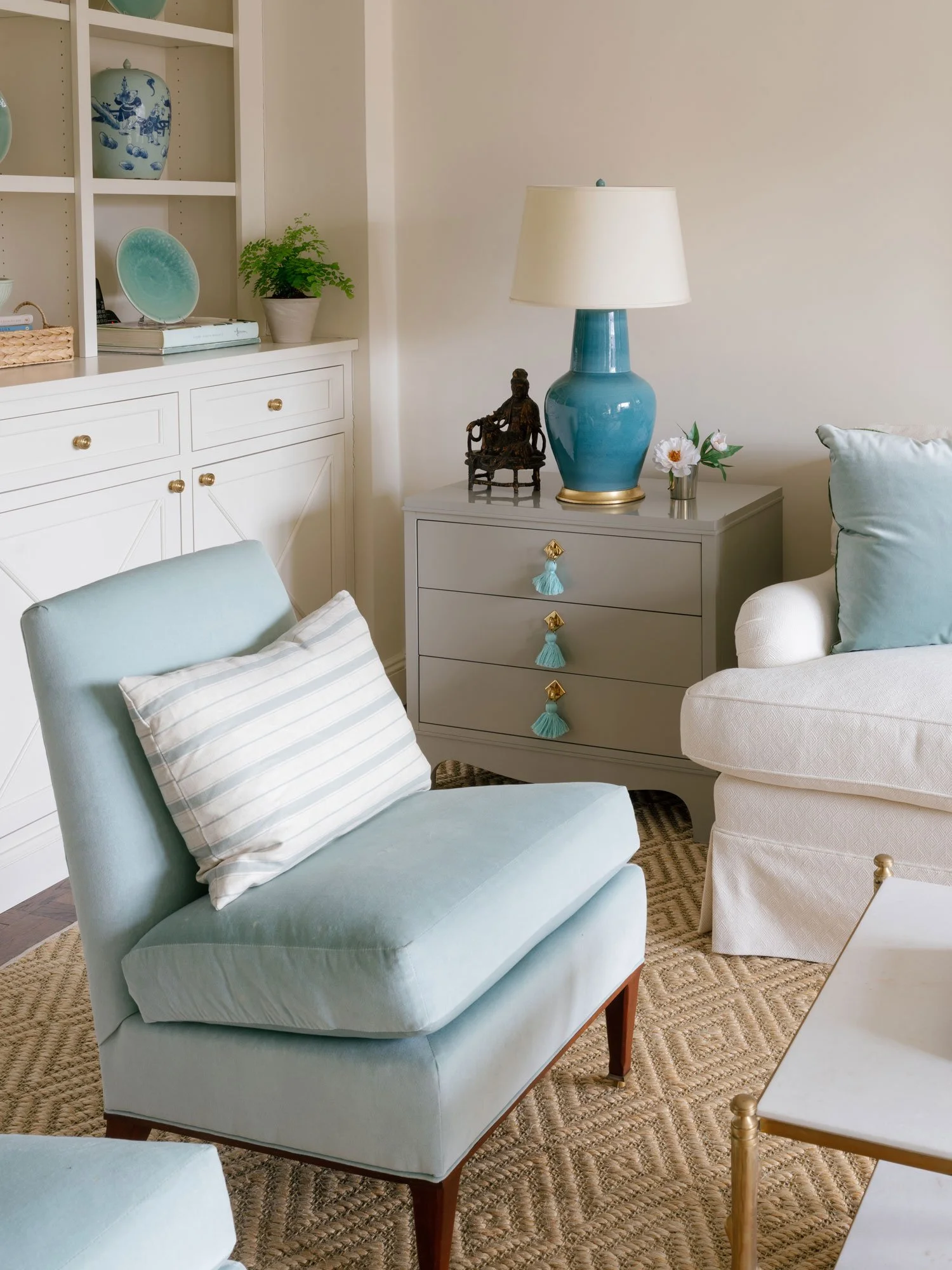 New York City apartment living room interior design by Elizabeth Hartz Interior Design featuring a pale blue accent chair, white upholstered seating, built-in shelving with collected ceramics, and layered neutral textures with soft blue accents.