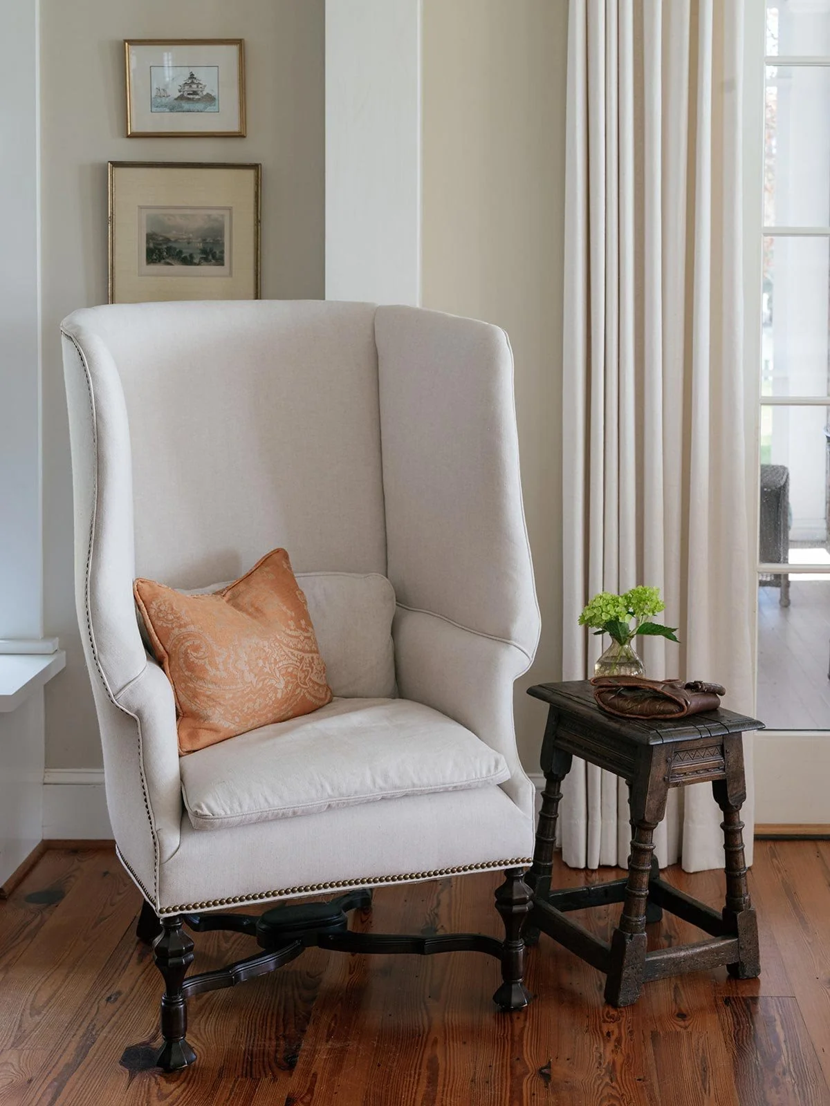 Cozy reading nook designed by Elizabeth Hartz Interior Design at The River home in Kilmarnock, Virginia. Features a high-back linen armchair with an orange accent pillow beside an antique wooden stool, layered with warm tones and natural light.