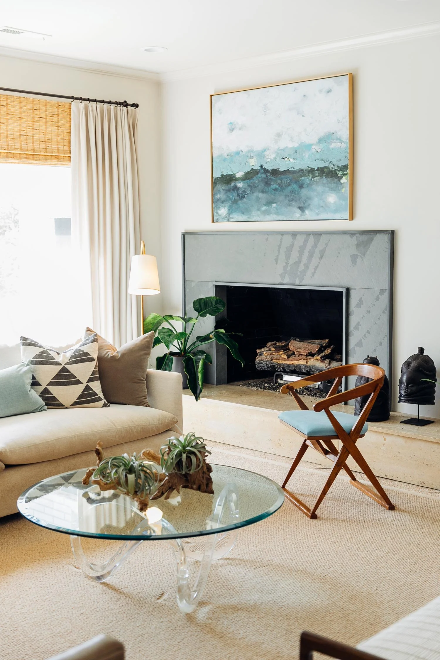 A cozy living room featuring a beige sofa with patterned pillows, a glass coffee table with decorative plants, a fireplace with logs, a wooden folding chair with a blue cushion, a potted plant, a modern art painting above the fireplace, and a window 