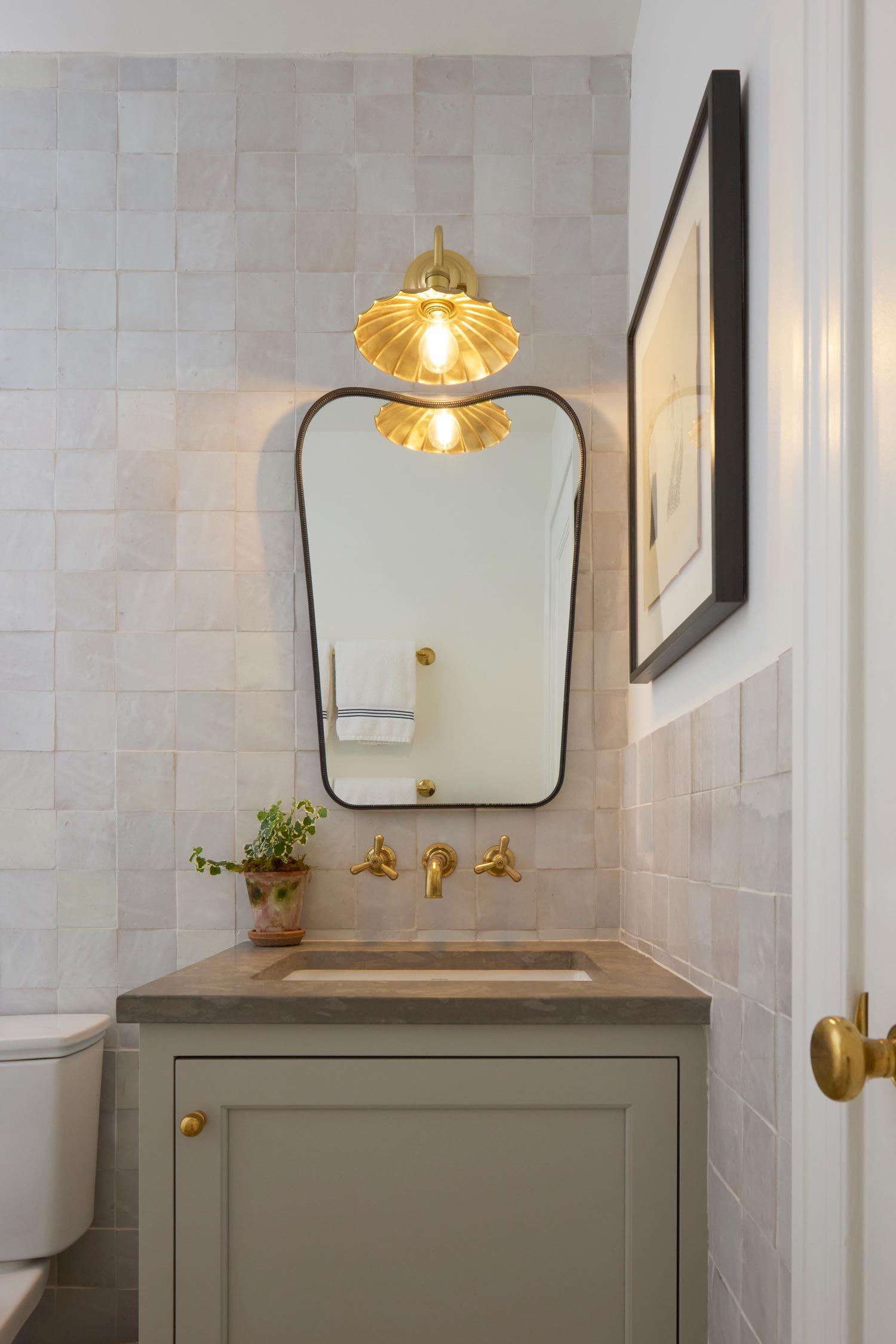 A small bathroom features a gray vanity with a framed mirror, gold fixtures, a potted plant on the counter, and a framed picture hanging on the wall. A white towel with black stripes is hanging on a gold towel bar reflected in the mirror. There is a 