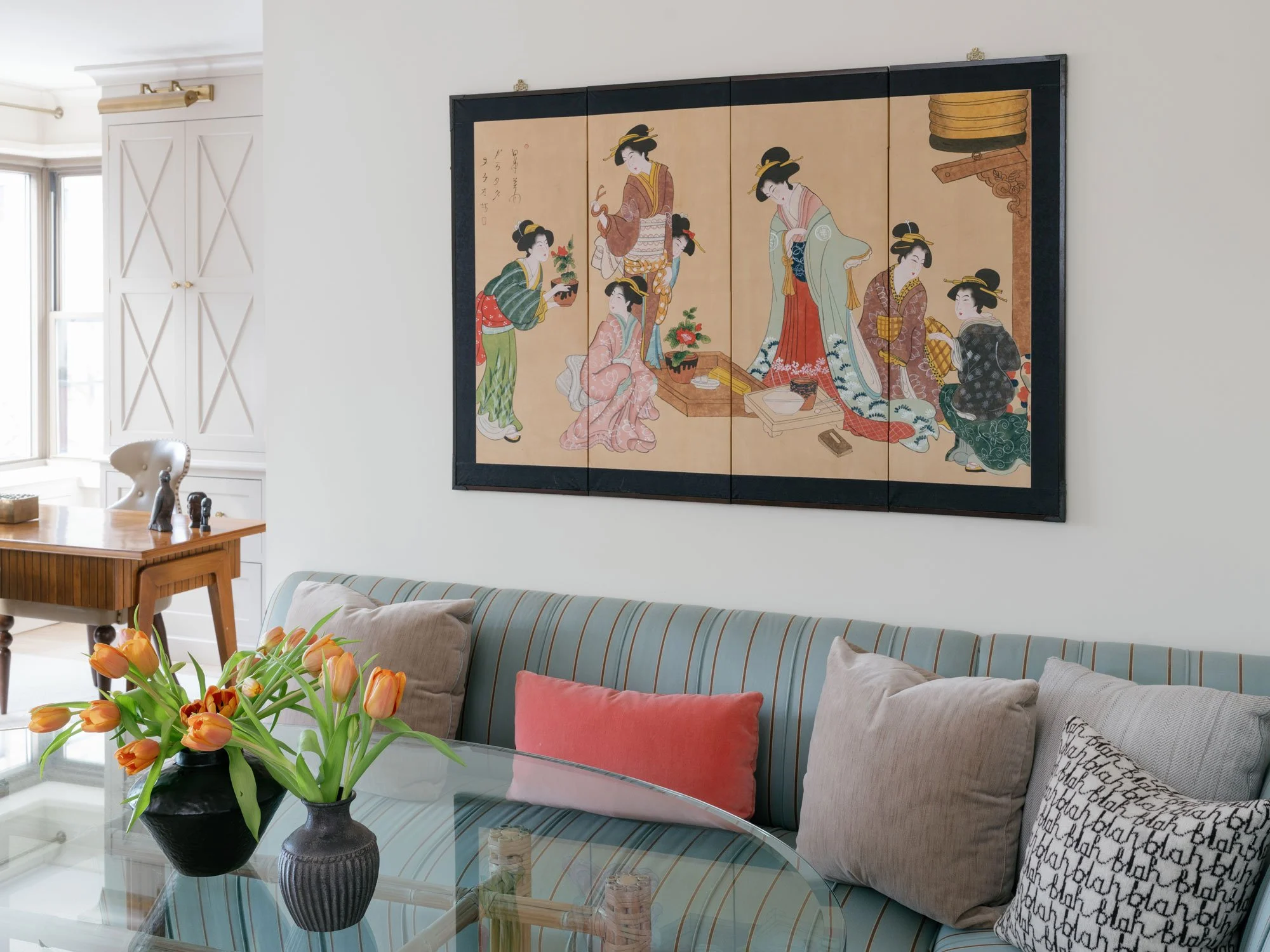 New York City apartment living room designed with a striped blue-green sofa, coral and neutral pillows, round glass coffee table, sculptural black vases with orange tulips, and statement artwork, showcasing layered, artful interior design.