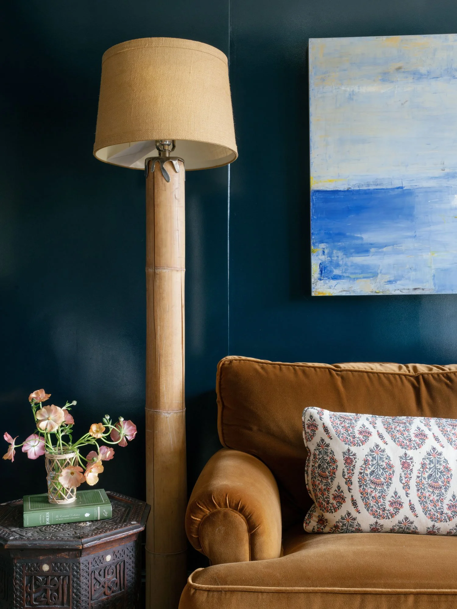 Cozy reading nook in a New York City apartment library featuring deep teal walls, bamboo floor lamp, carved wood side table with flowers and books, and a plush camel-toned velvet sofa styled with a patterned accent pillow.