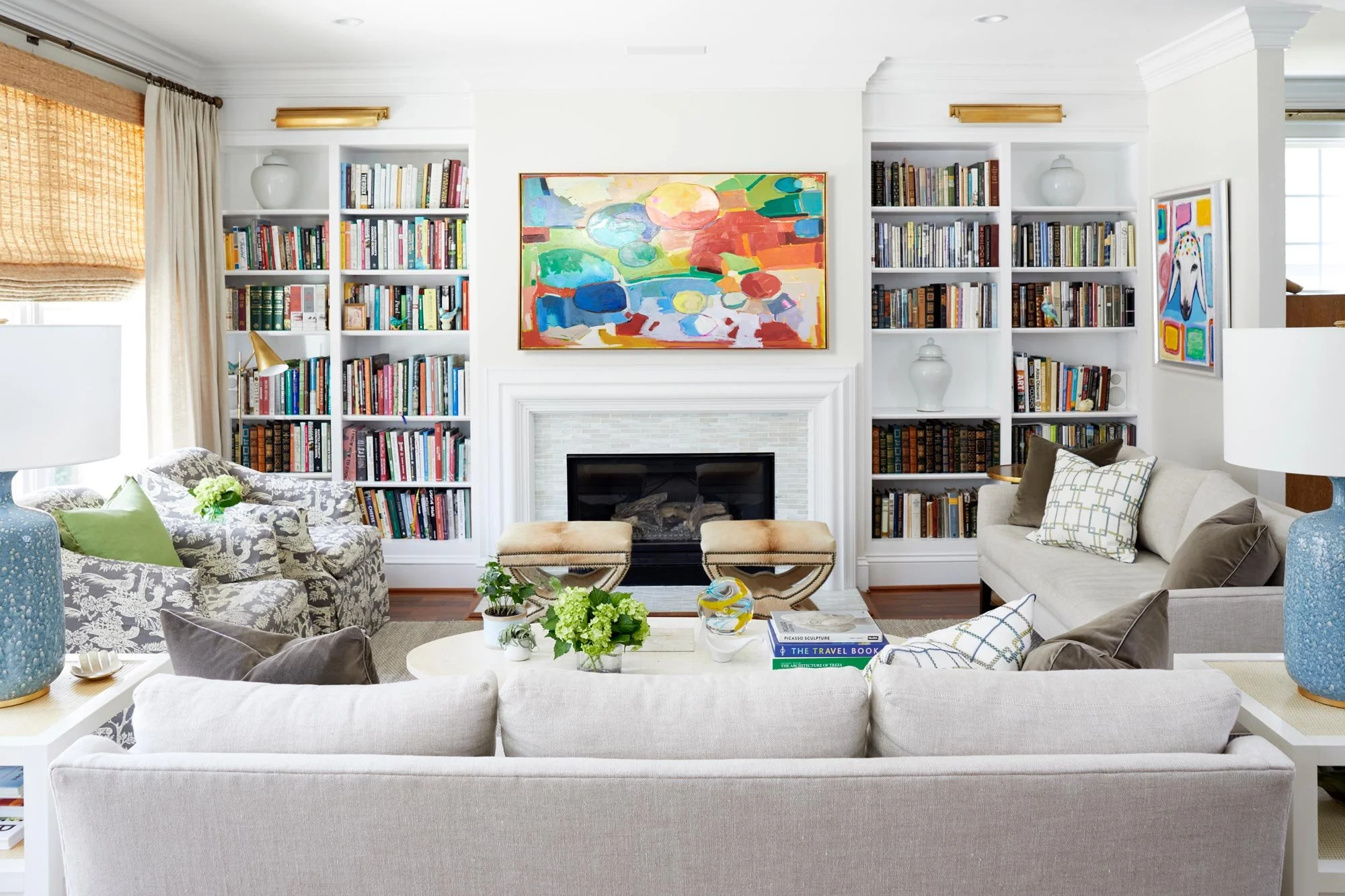 Living room with white furniture, built-in bookshelves filled with books, colorful painting above fireplace, two beige chairs, patterned armchair, coffee table with plants, books and decorative items, lamps, windows with curtains.