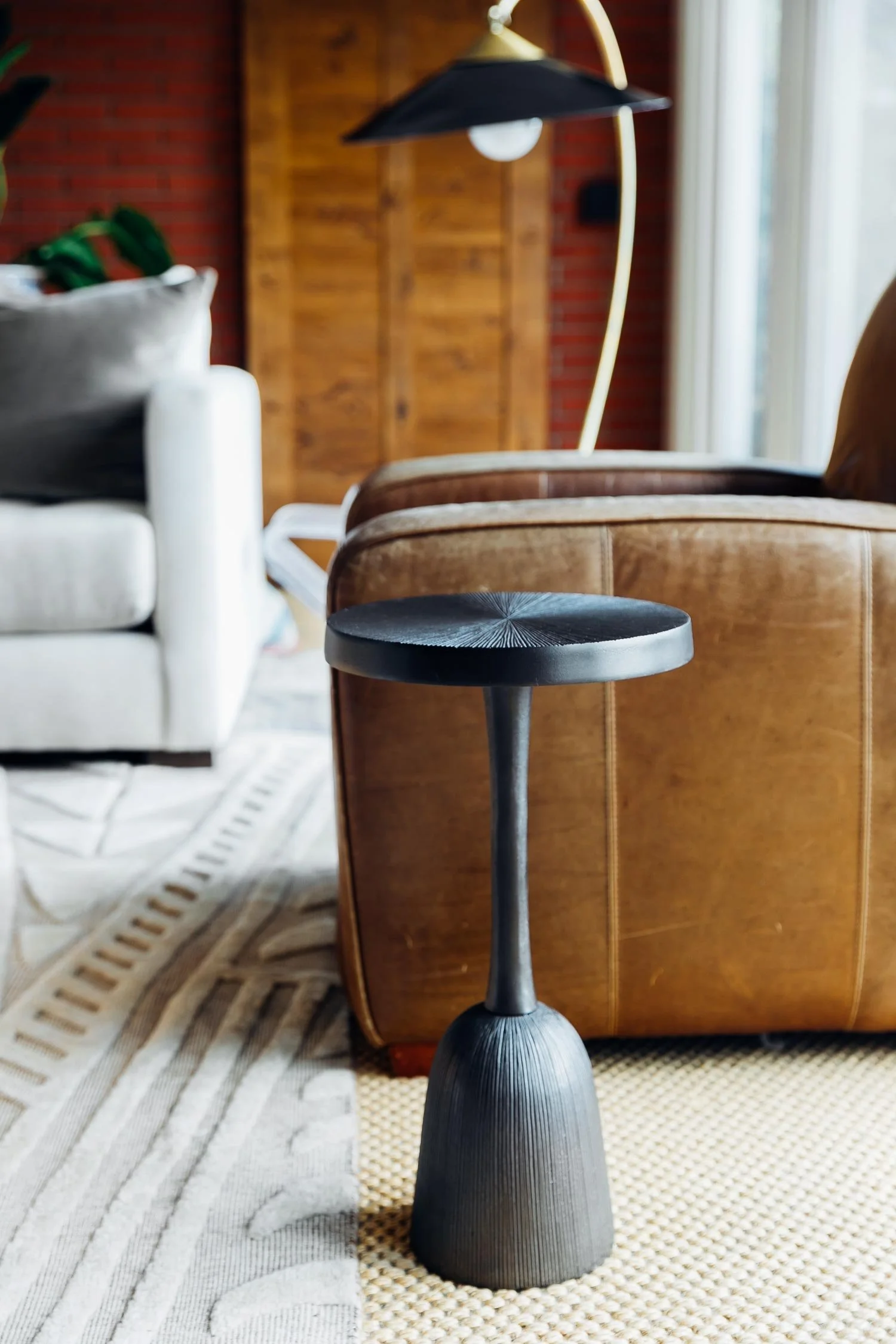 Black modern side table standing on a beige woven rug in a cozy living room with a leather armchair, white sofa, wooden wall, and large window with curtains.