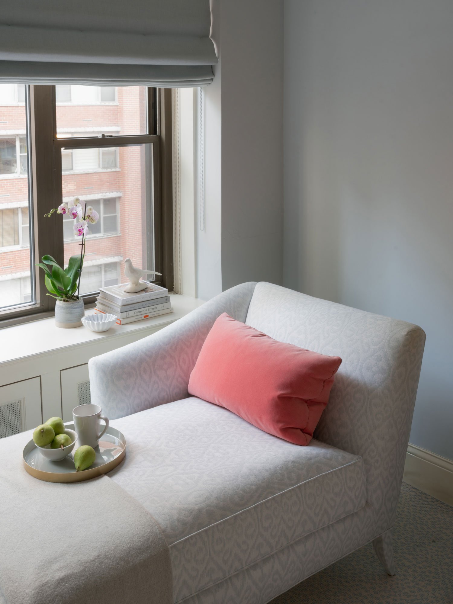 New York City bedroom seating nook by Elizabeth Hartz Interior Design featuring upholstered chaise lounge, coral accent pillow, styled window ledge with orchids, and soft layered textiles