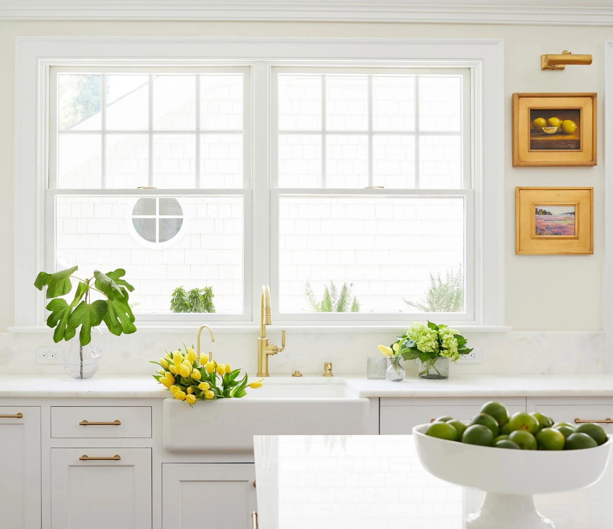 Bright kitchen with white cabinets, large window, and yellow tulips on the counter. There are potted plants, artworks on the wall, and a bowl of lemons outside the window.