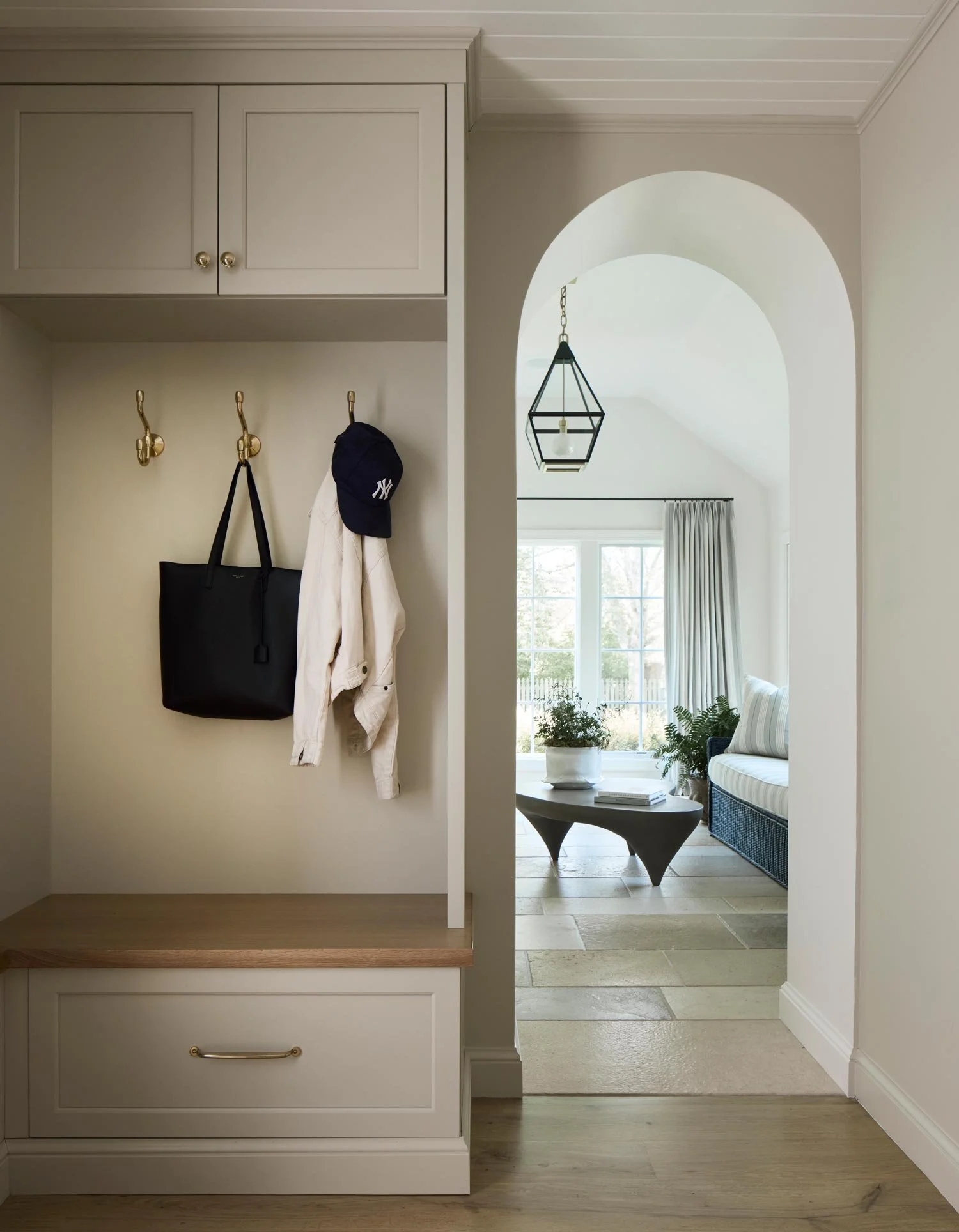 Entryway with hooks holding a black bag, white jacket, and a dark blue cap, next to an archway leading to a living room with a large window, plants, and modern furniture.