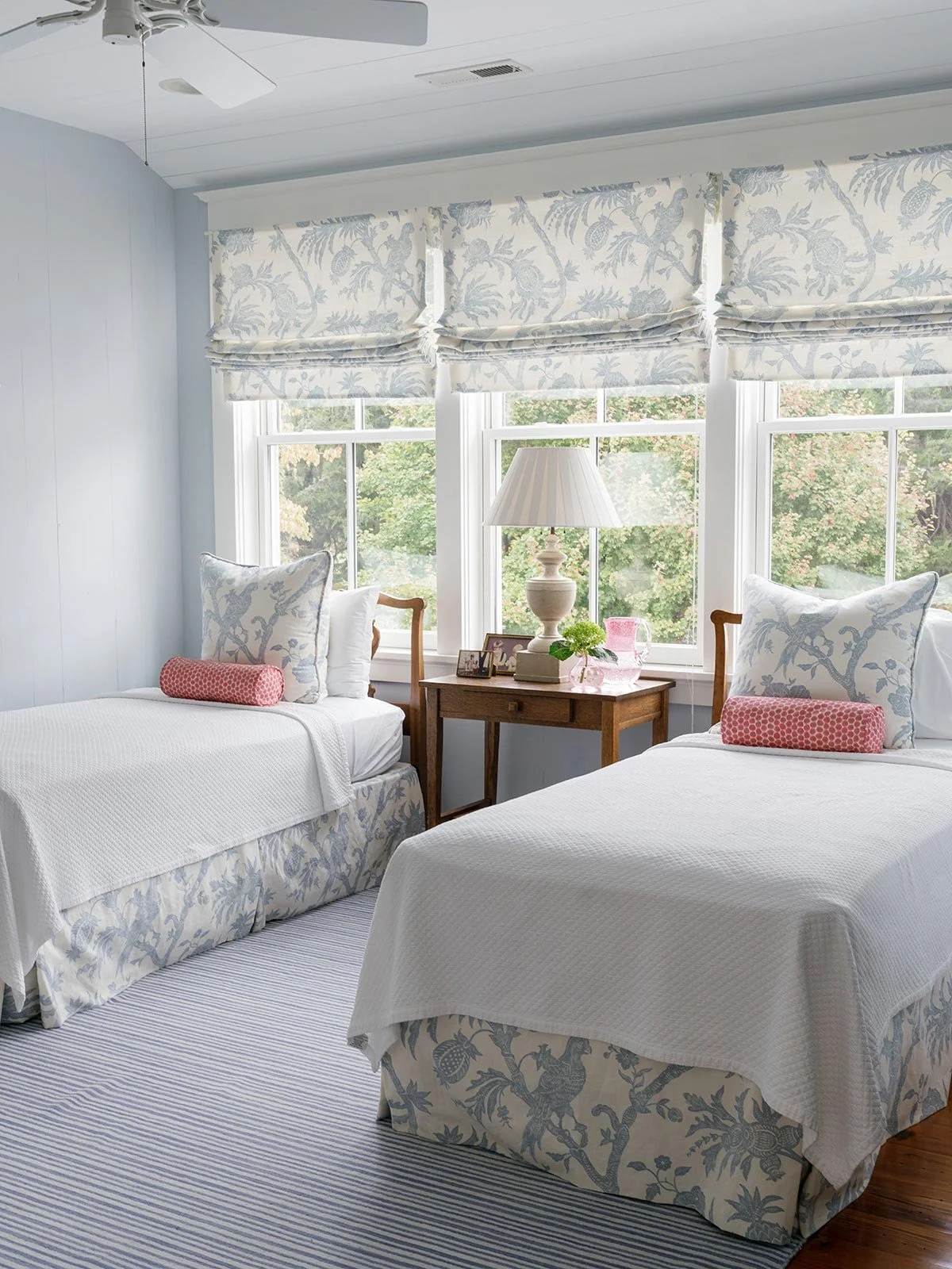 Light and airy twin bedroom designed by Elizabeth Hartz Interior Design at The River home in Kilmarnock, Virginia. Features matching beds with blue and white botanical fabrics, striped rug, and soft Roman shades, creating a coastal-inspired guest roo