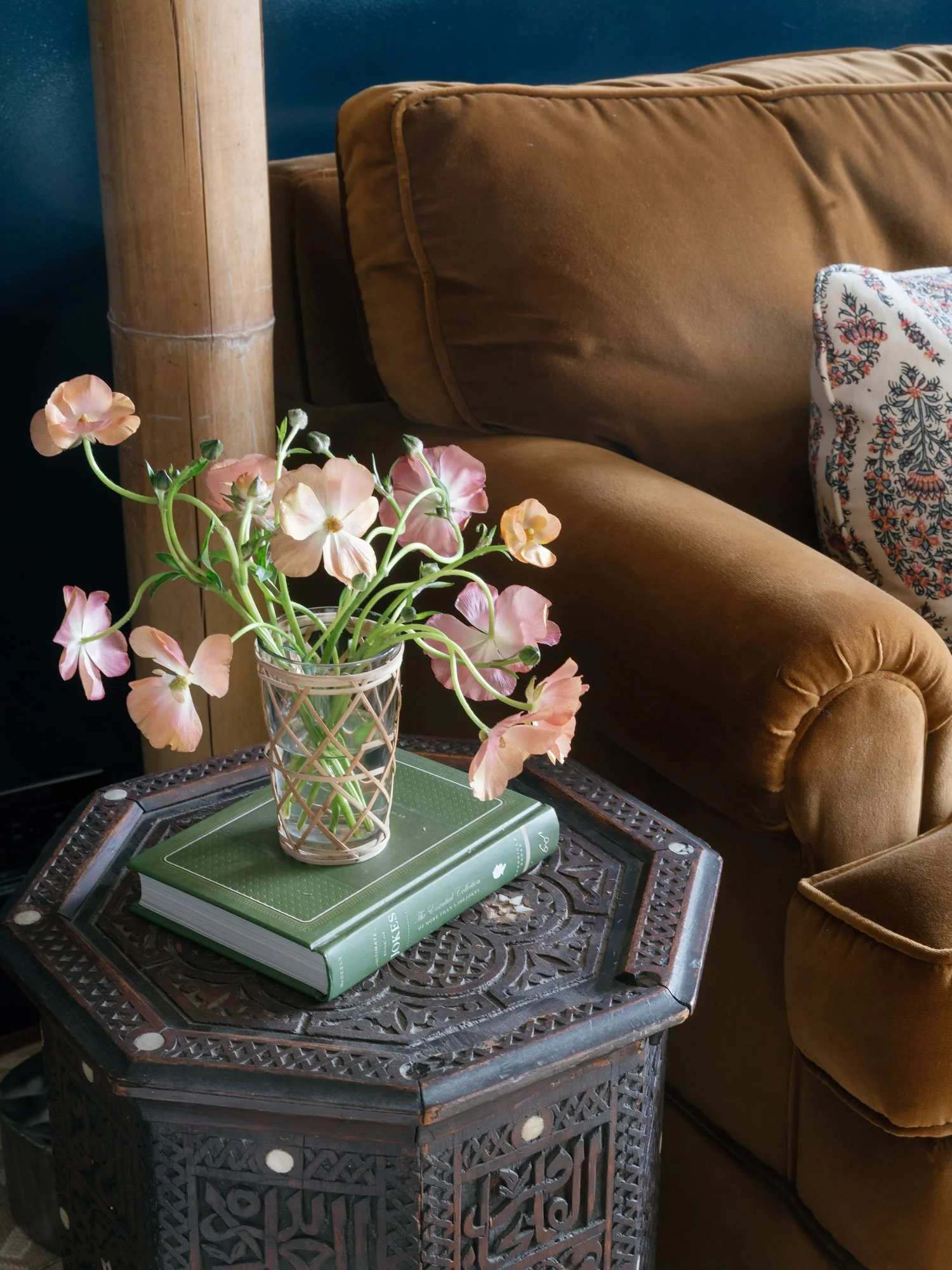 Detail of library TV room in New York City apartment by Elizabeth Hartz Interior Design, featuring caramel velvet sofa, dark blue wall, carved wood side table, and soft pink flowers styled on books for a warm, layered seating vignette.