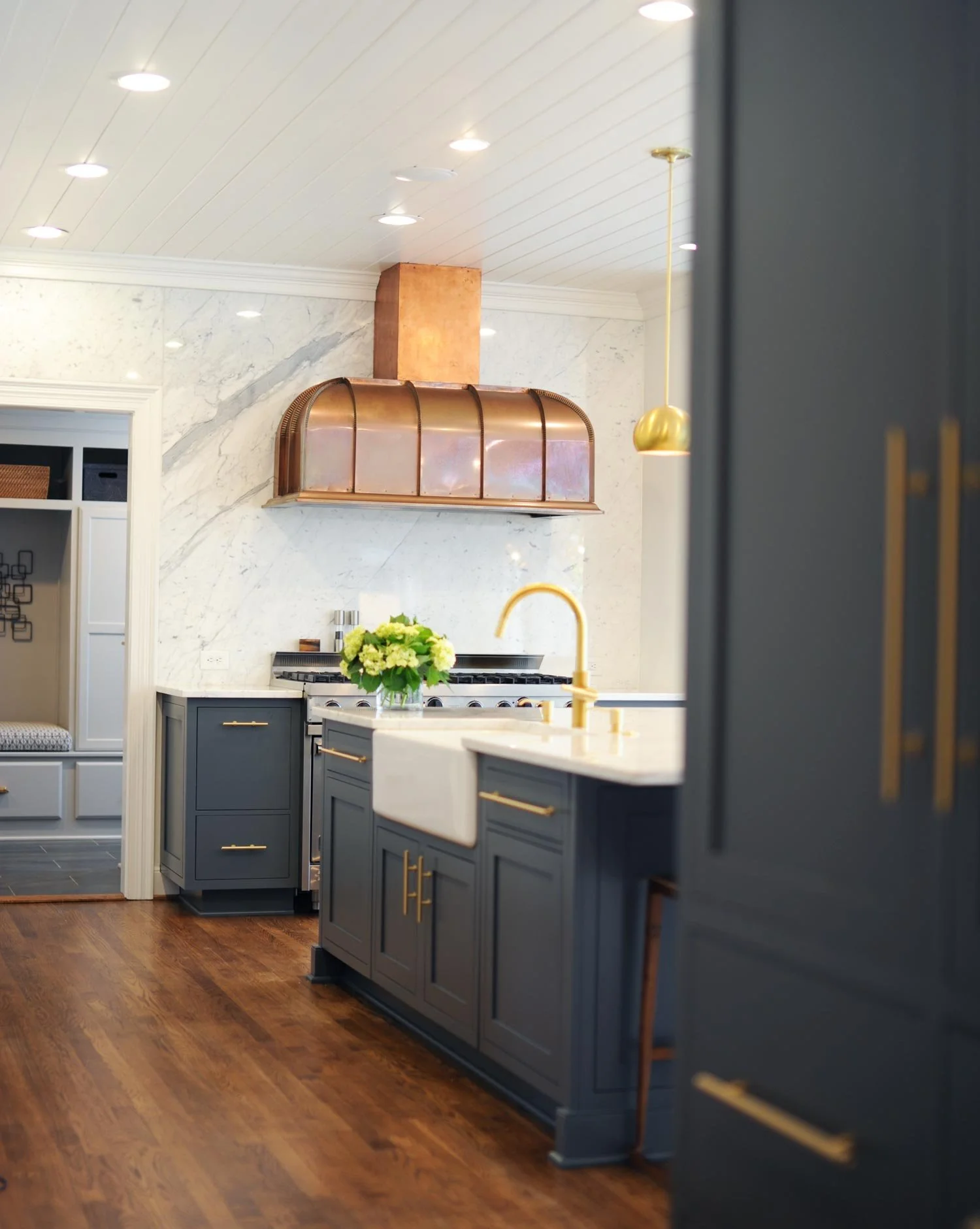 Elizabeth Hartz designed a modern kitchen with navy cabinets, gold hardware, marble backsplash, copper range hood, wooden floors, and a white farmhouse sink.
