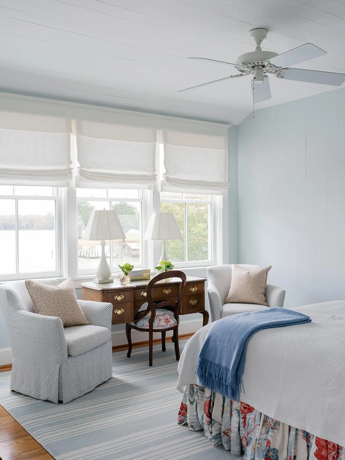Serene bedroom designed by Elizabeth Hartz Interior Design at The River home in Kilmarnock, Virginia. Features soft blue walls, striped area rug, antique wooden desk, and upholstered chairs beside large windows, blending coastal calm with traditional