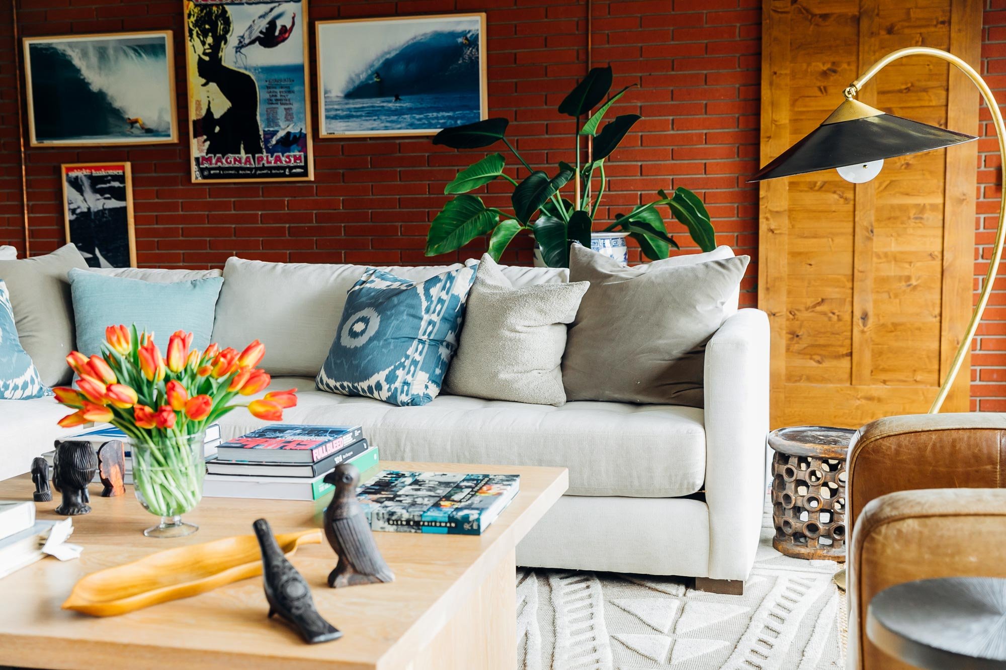 Living room with a beige sofa decorated with blue and neutral pillows, a wooden coffee table holding a vase of red and yellow tulips, stacked books, and decorative animal sculptures. A large green leafy plant is behind the sofa, and a wooden door and