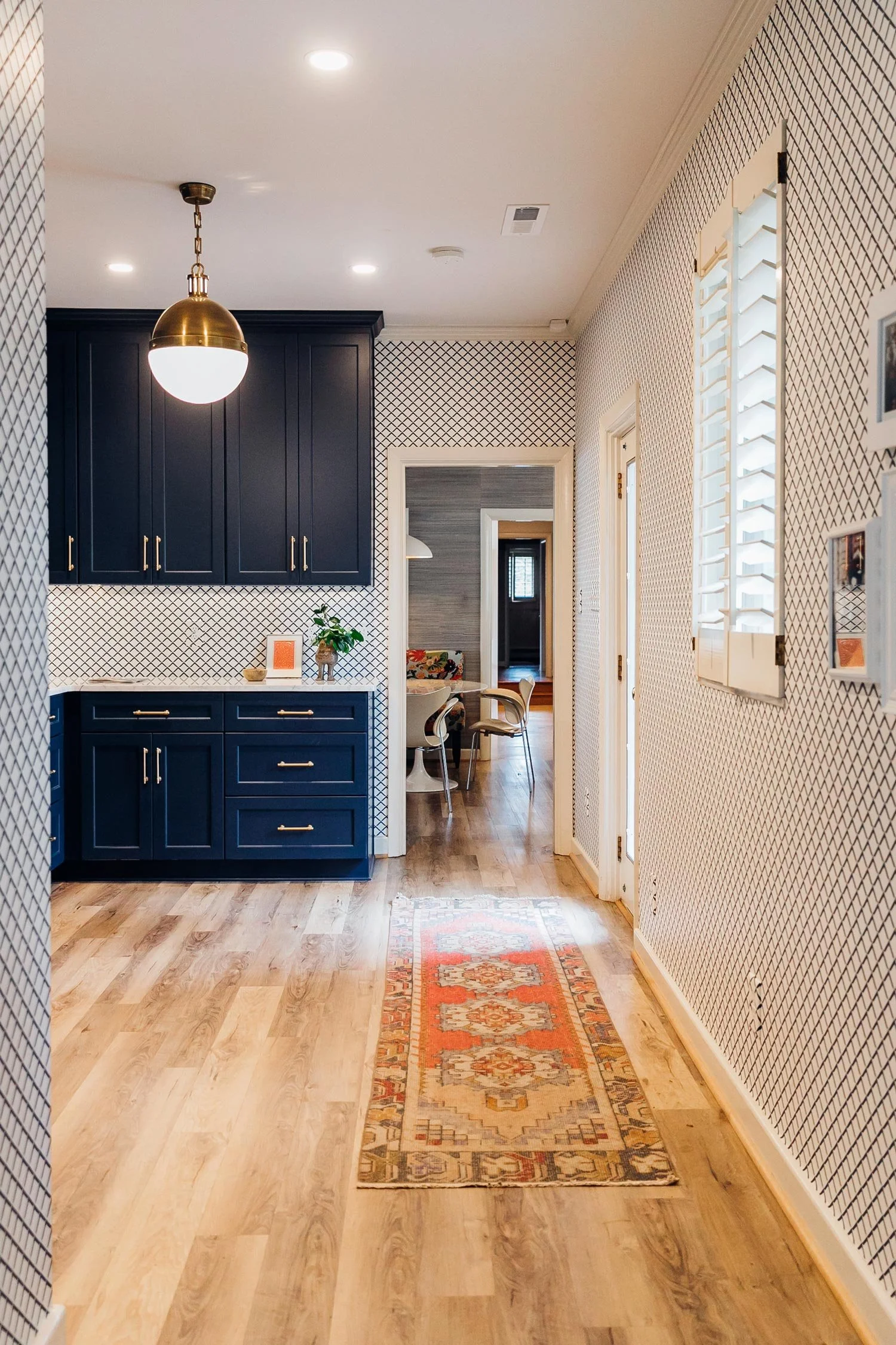 View of a hallway with wooden floor, patterned wallpaper, a window with white plantation shutters, a ceiling light, a dark blue cabinet, a small decorative rug, and a doorway leading to a dining area with chairs and a table.