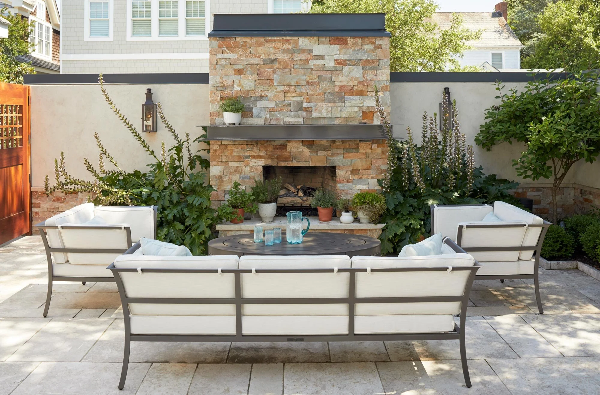 Outdoor patio with white cushioned seating arranged around a round coffee table, in front of a stone fireplace decorated with potted plants, against a wall with outdoor lanterns, surrounded by greenery and trees.