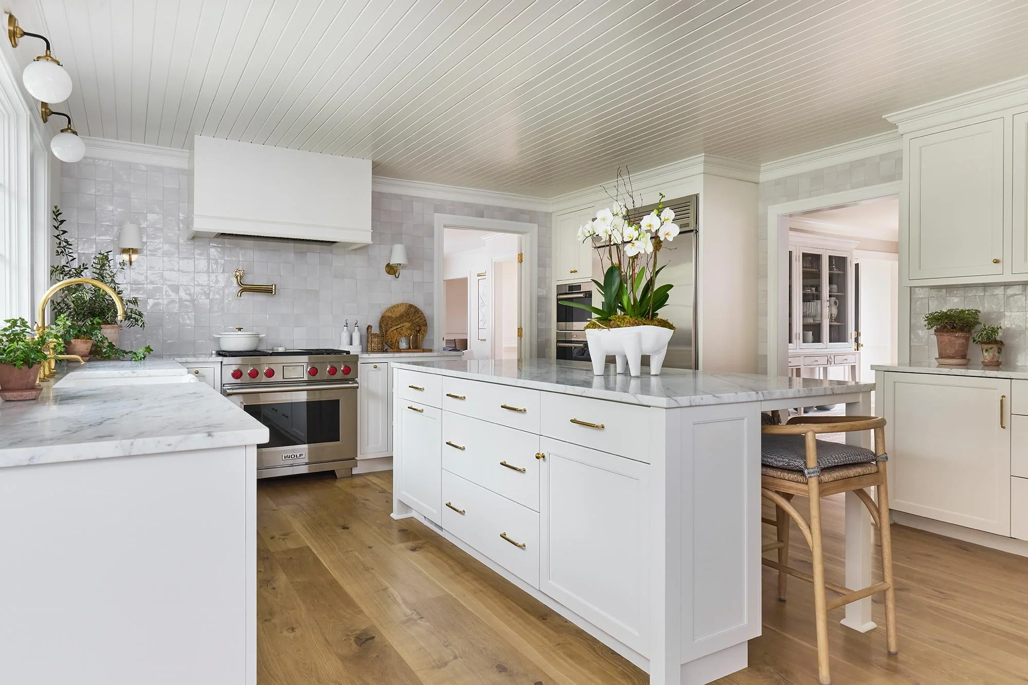 White kitchen with marble countertops, gold hardware, potted plants, orchid centerpiece on island, wooden stool, and modern appliances.