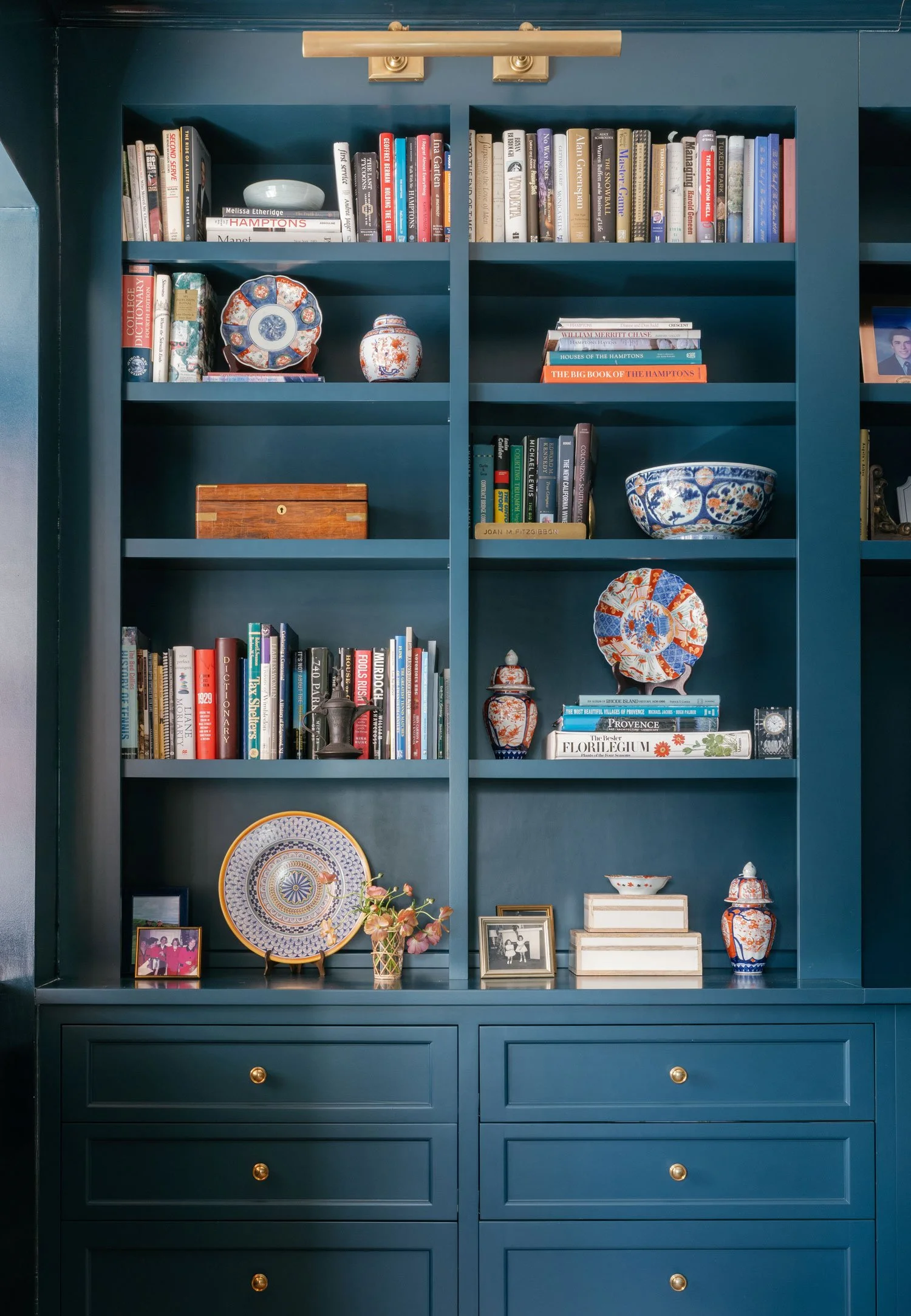 Bold blue built-in bookcases in New York City apartment library TV room designed by Elizabeth Hartz Interior Design, styled with collected books, blue and white porcelain, framed photos, and brass picture light for a layered, tailored look.