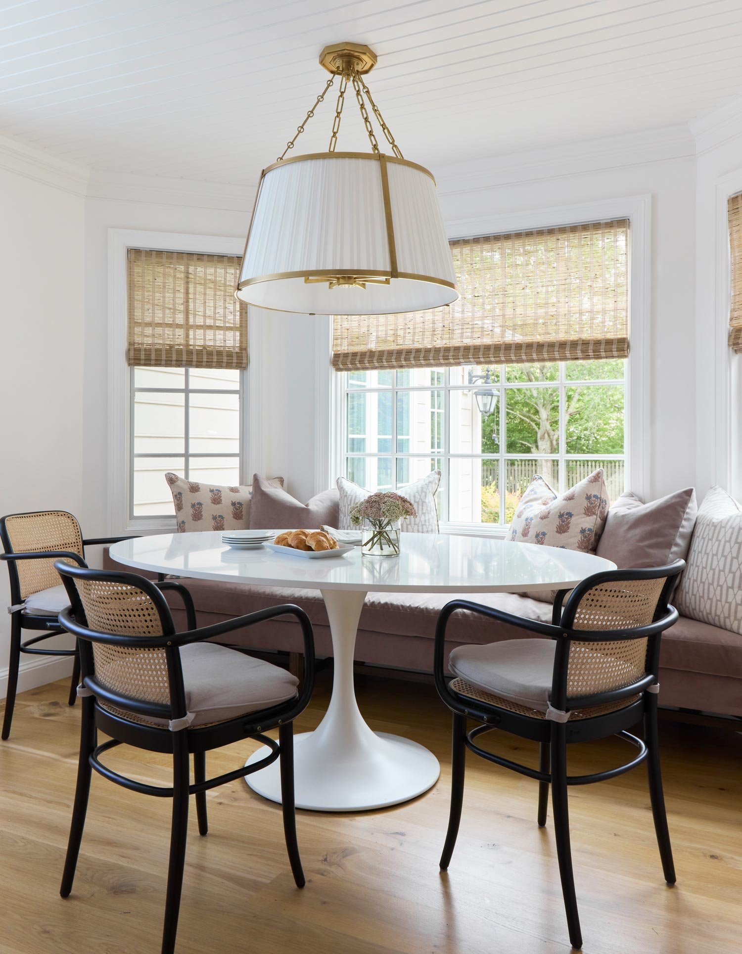 Bright dining nook with a white round table, black armchairs, and plush corner cushions near large windows with bamboo blinds, topped by a white drum chandelier.