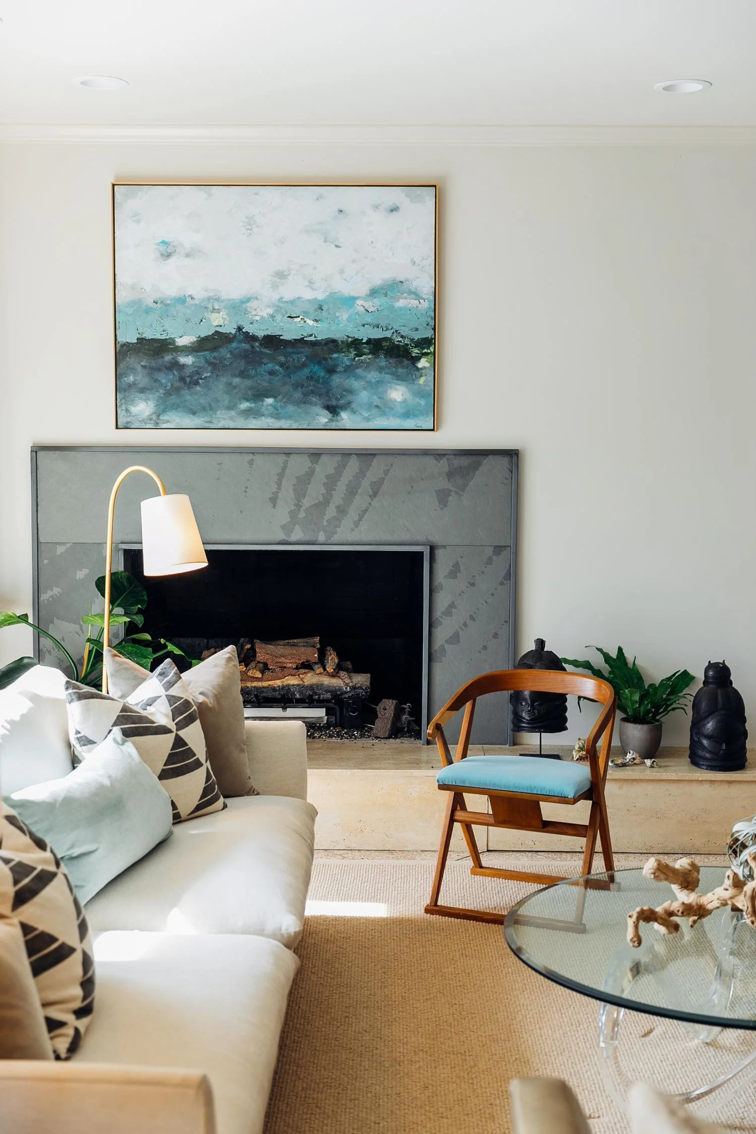 A living room with a white sofa, decorative pillows, a glass coffee table, a blue armchair, a modern fireplace, a large abstract painting, potted plants, and decorative black sculptures.