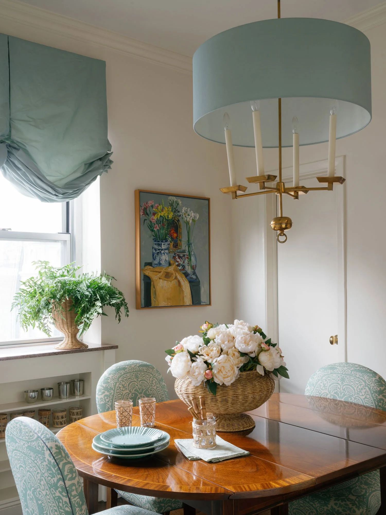 Dining room in New York City apartment designed by Elizabeth Hartz Interior Design featuring aqua patterned upholstered dining chairs, polished wood dining table, woven floral centerpiece, soft blue Roman shade, and brass chandelier with custom blue 