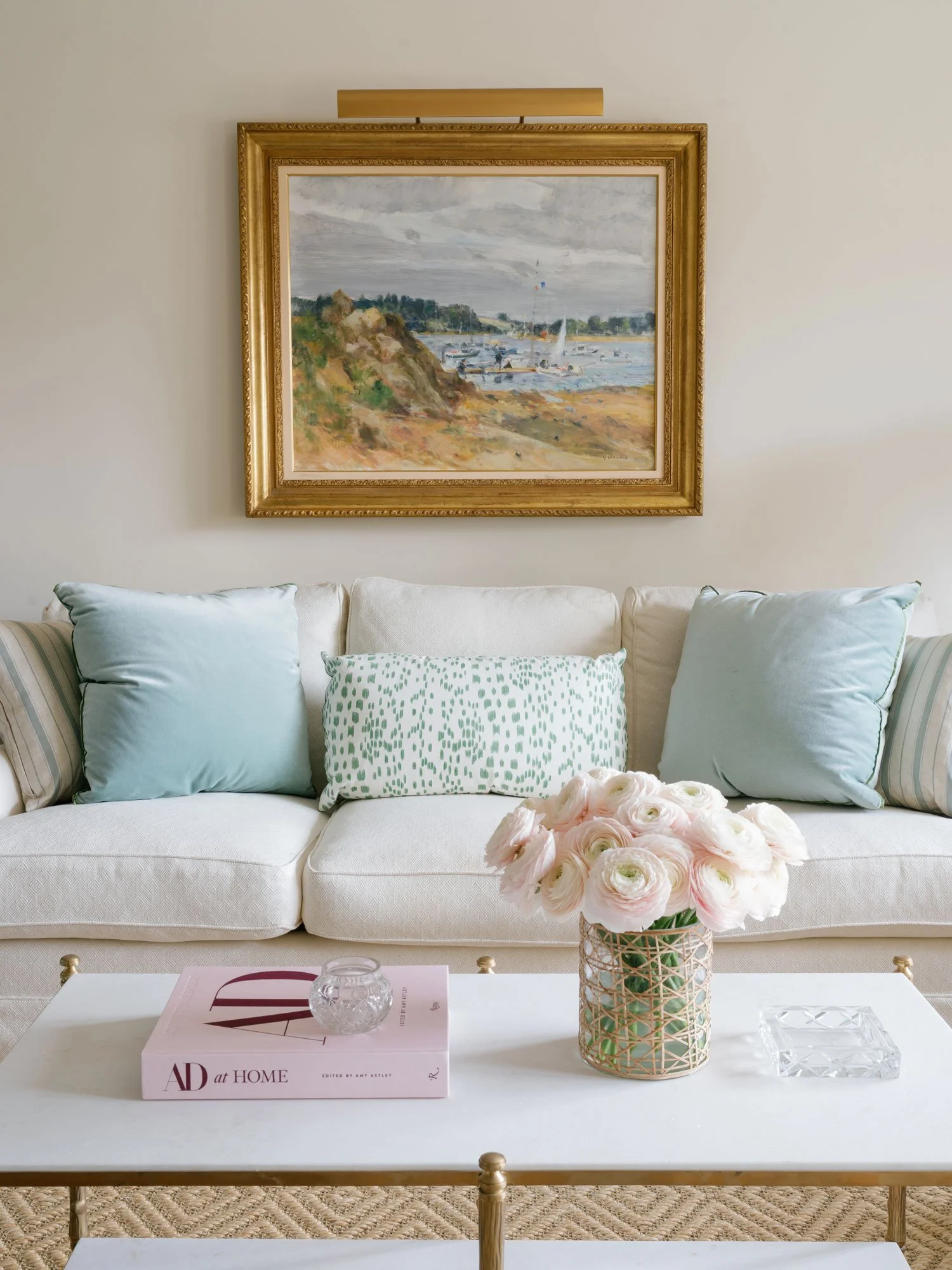 New York City apartment living room interior design by Elizabeth Hartz Interior Design featuring a neutral sofa with soft blue and patterned accent pillows, layered coffee table styling, fresh florals, and classic artwork above the sofa.