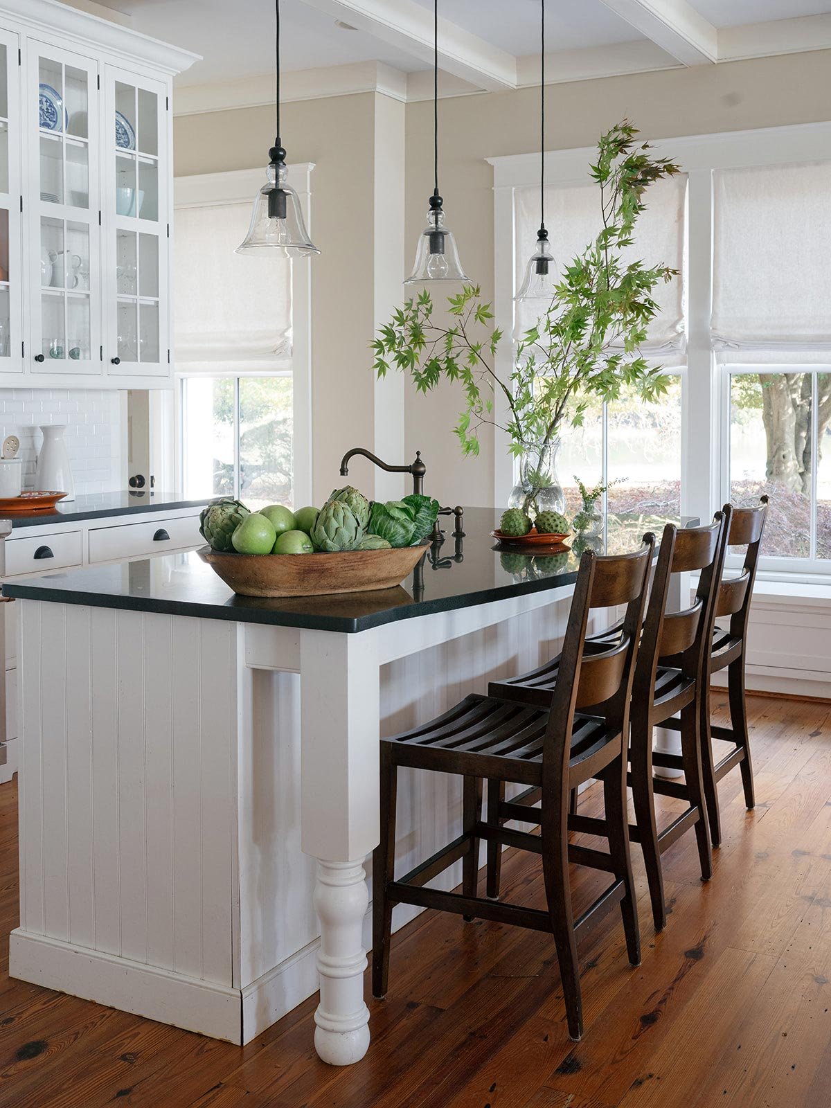 Coastal kitchen designed by Elizabeth Hartz Interior Design at The River home in Kilmarnock, Virginia. Features a white island with dark stone countertop, wooden stools, glass pendant lights, and fresh greenery for a bright, timeless coastal look.