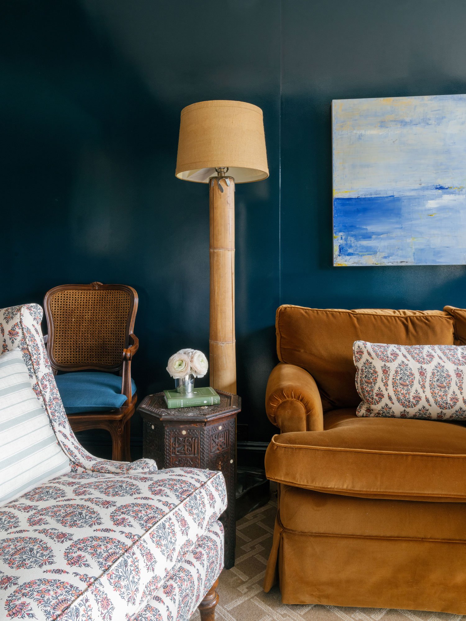 Library TV room in New York City apartment by Elizabeth Hartz Interior Design featuring deep blue walls, caramel velvet sofa, patterned accent chair, cane-back chair, bamboo floor lamp, and layered textiles creating a cozy, collected seating area.