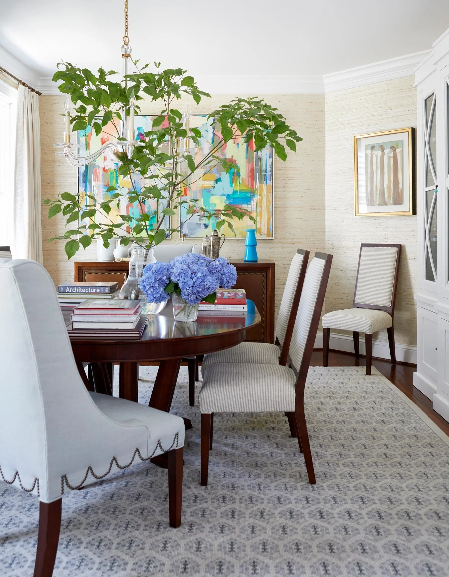 A dining room with a wooden table and upholstered chairs, decorated with a large potted plant with green leaves and blue hydrangea flowers, a colorful abstract painting on the wall, and a white cabinet. The room has a patterned rug and cream-colored 