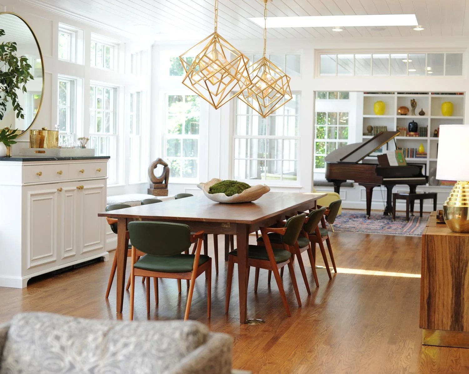 Bright living and dining room in Virginia Beach with large windows, wooden flooring, modern chandelier, wooden dining table with green chairs, white sideboard, and a grand piano.