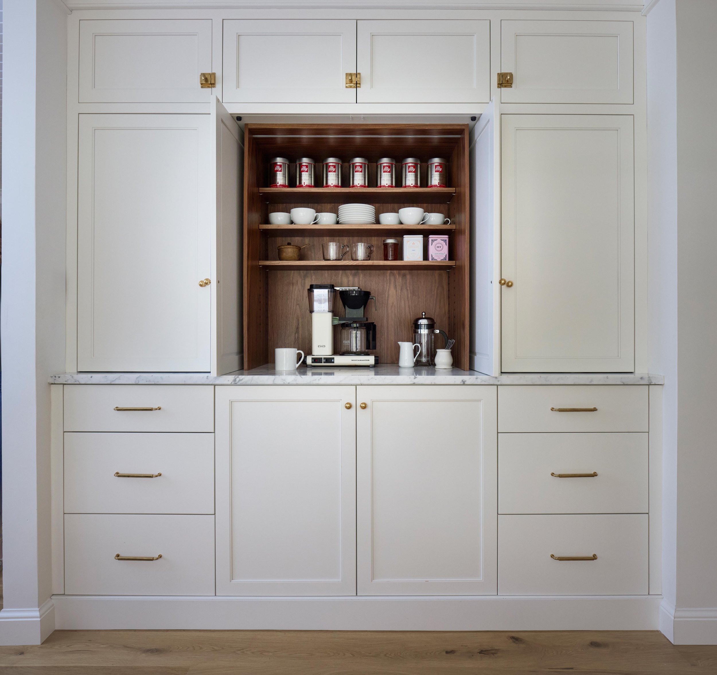 White kitchen cabinet with opened middle section showing shelves with coffee supplies, cups, and jars, and small appliances on the counter, with gold-colored handles.