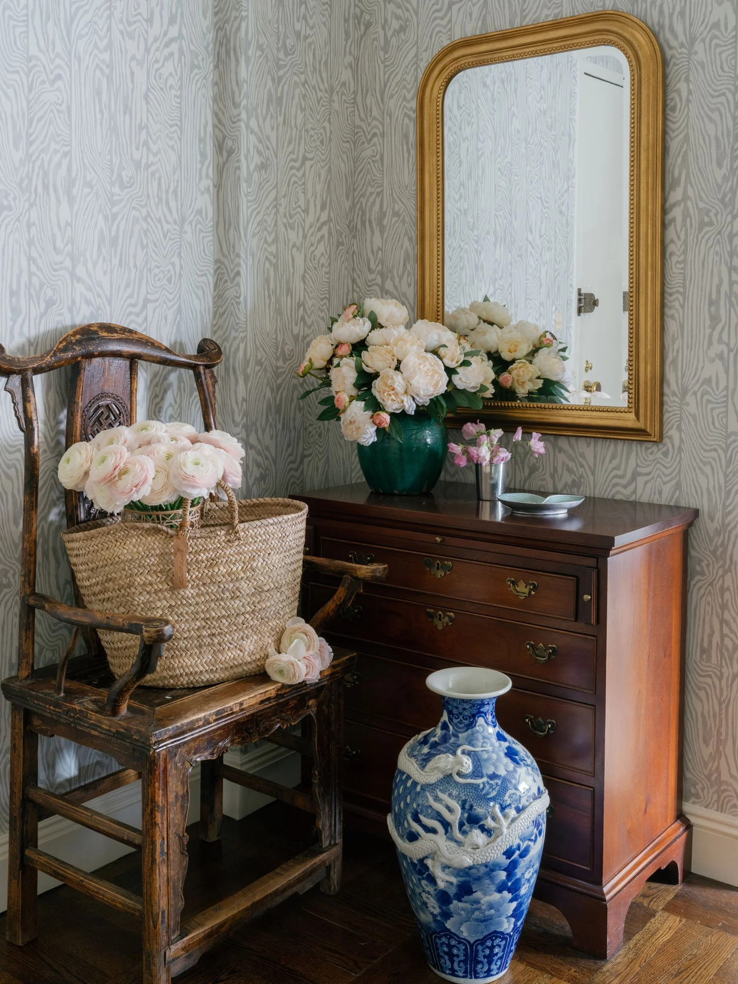 Entryway of New York City apartment designed by Elizabeth Hartz Interior Design featuring antique wooden chair, woven basket with pale blush flowers, dark wood chest, gold framed mirror, blue and white porcelain floor vase, and layered floral styling