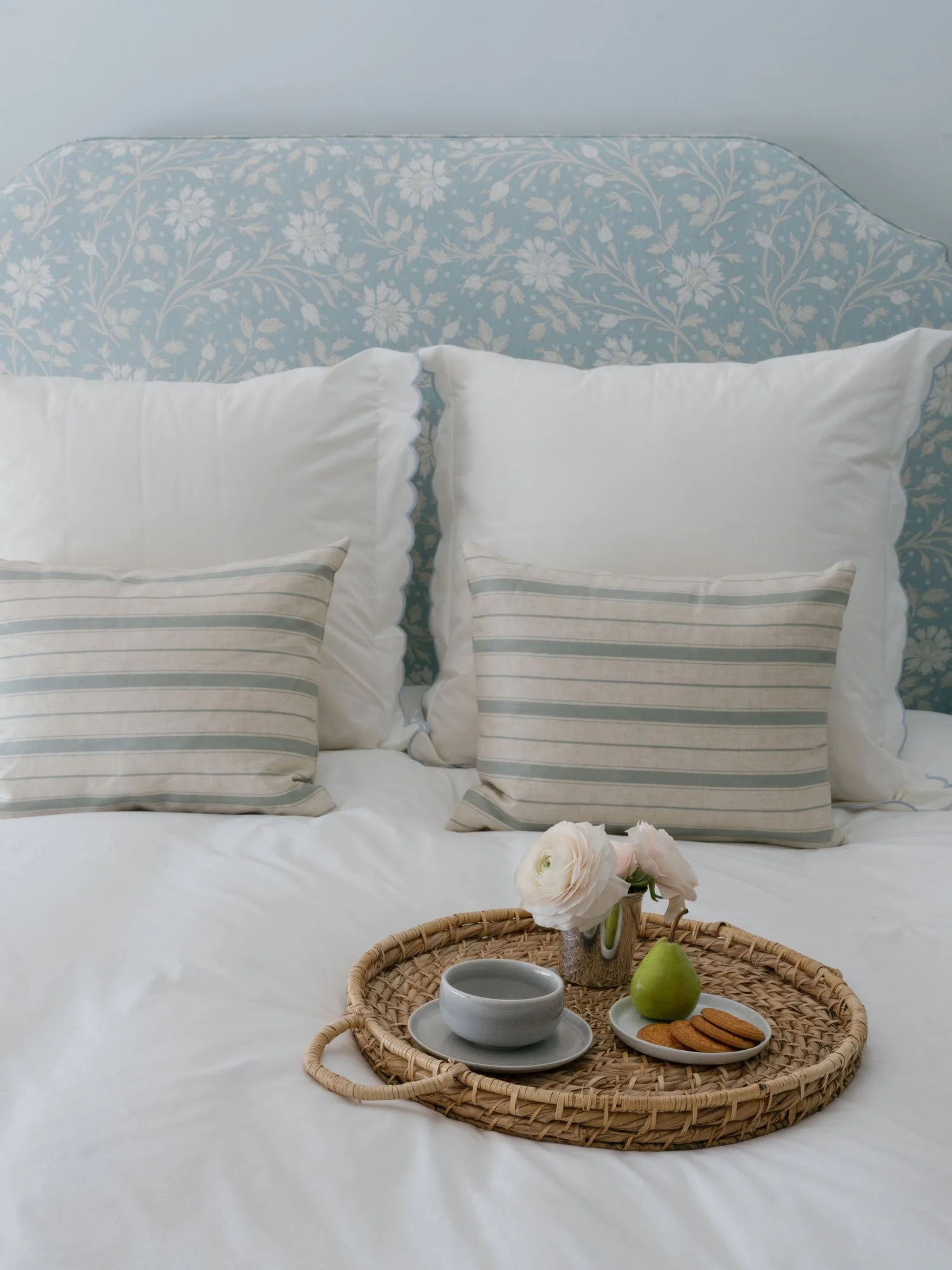 Serene New York City apartment bedroom featuring a pale blue floral upholstered headboard, crisp white bedding with striped accent pillows, and a woven tray on the bed styled with coffee, fresh flowers, a pear, and cookies.