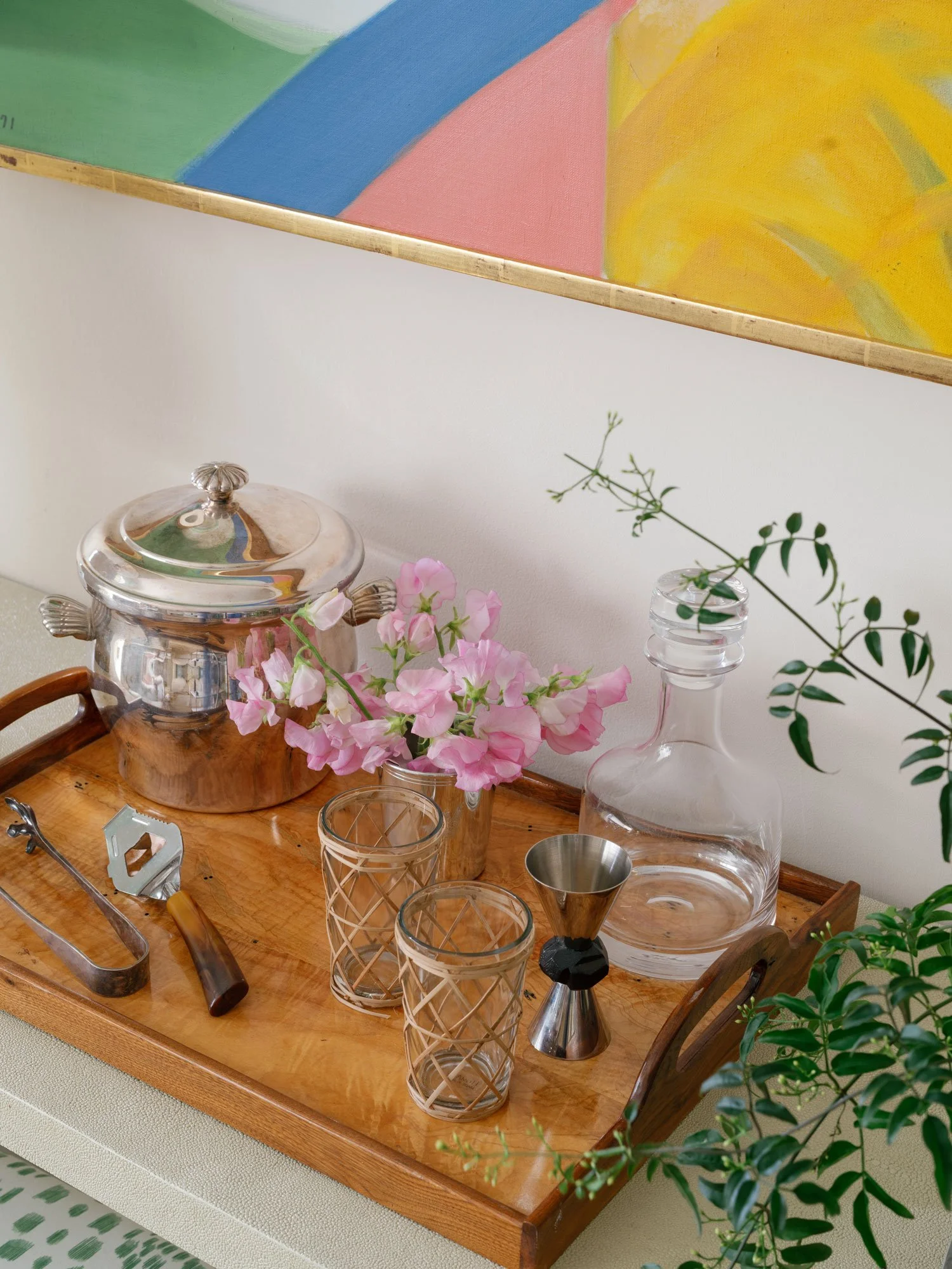 Styled bar tray detail in New York City apartment by Elizabeth Hartz Interior Design featuring crystal decanter, vintage ice bucket, glassware, cocktail tools, and fresh florals layered beneath colorful abstract artwork for a refined, southern-inspir