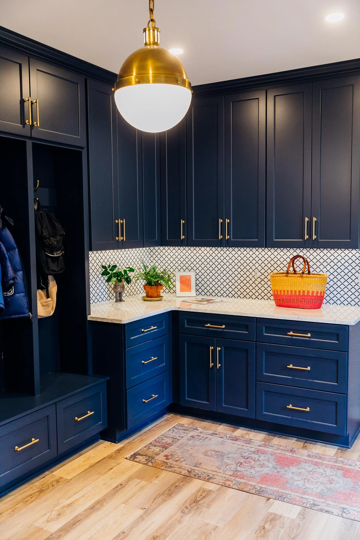 A modern foyer with navy blue cabinetry, gold handles, and a white quartz countertop. The area includes a corner with plants, a framed picture, and a colorful woven tote bag. A decorative rug is on light wood flooring, and a gold pendant light hangs 