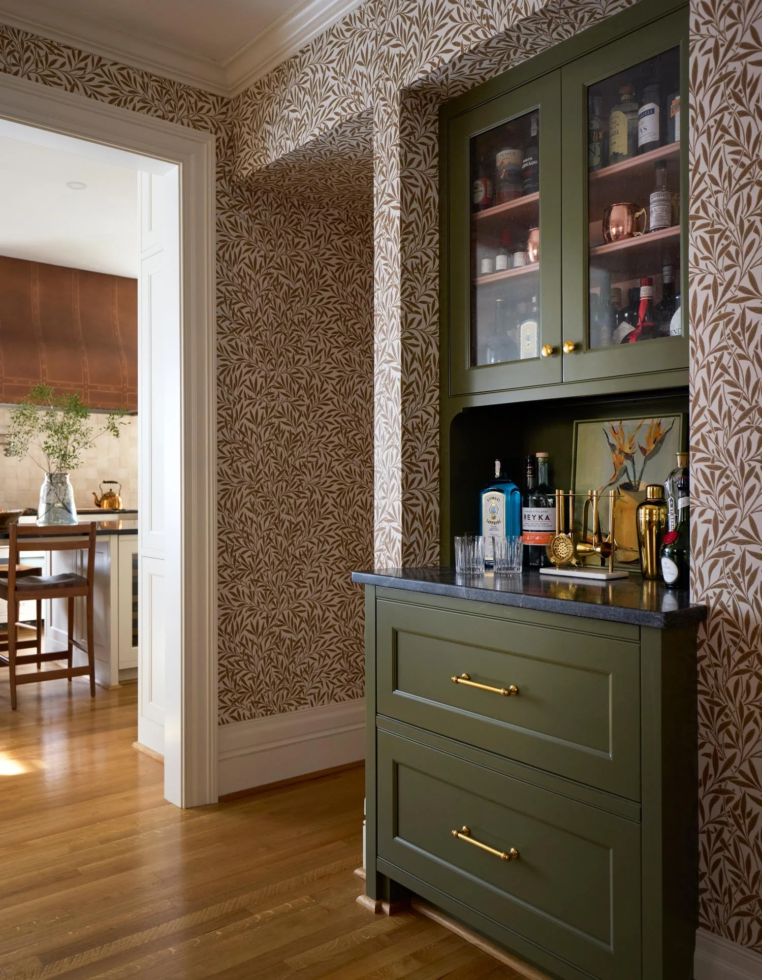 A green bar cabinet with glass doors and gold handles, displaying bottles of alcohol, shot glasses, and decorative items in a room with patterned wallpaper and wooden flooring.