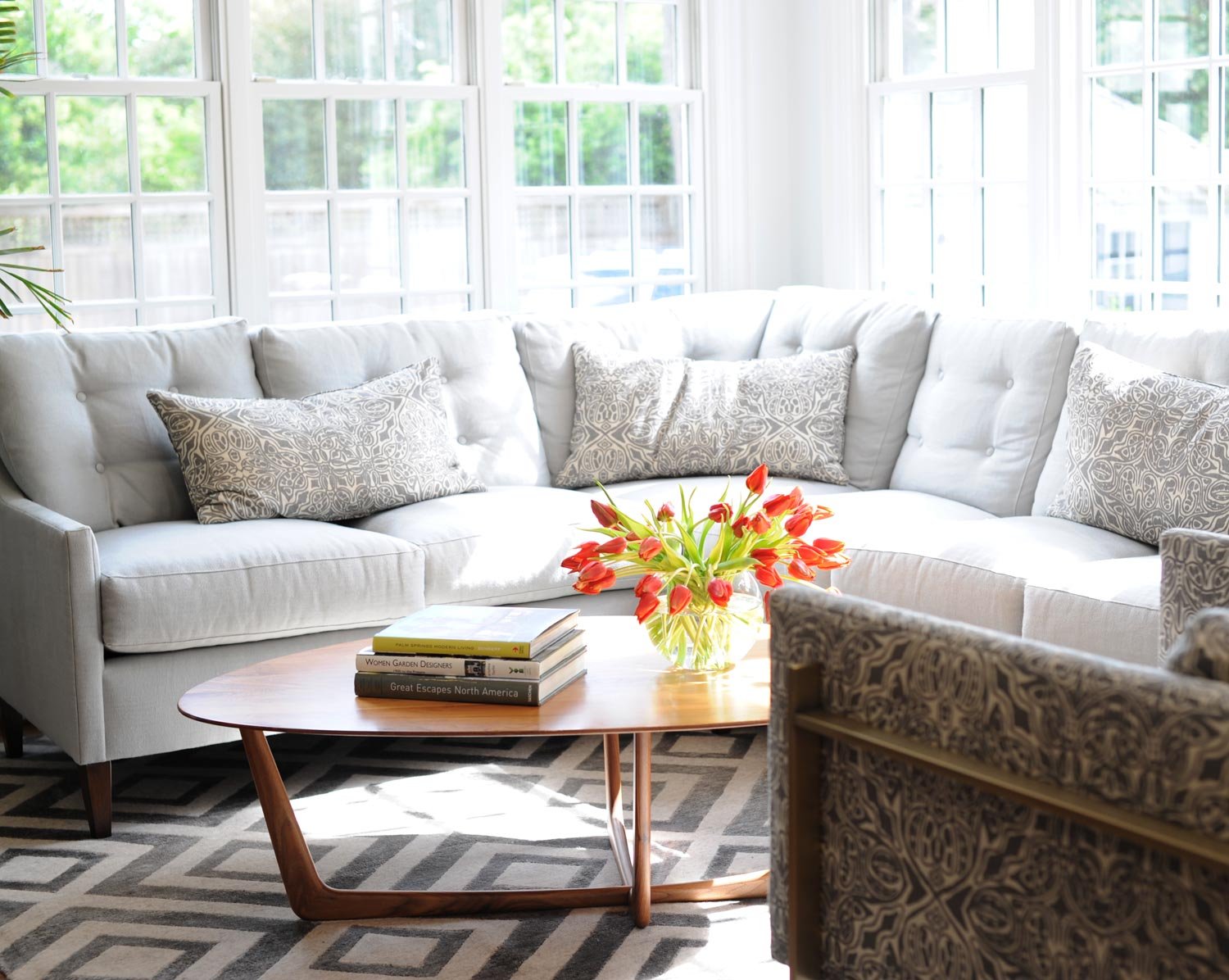 Bright living room in Virginia Beach with a white sectional sofa, patterned throw pillows, a wooden coffee table with books and a vase of red tulips, large windows with white frames letting in natural light, and a patterned area rug.