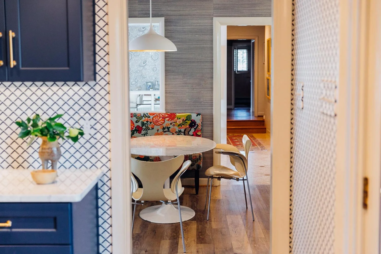 View through doorway to a dining area with a marble table, floral print seating, and cream chairs. Wooden flooring, wall decor, and a hanging lamp are also visible.