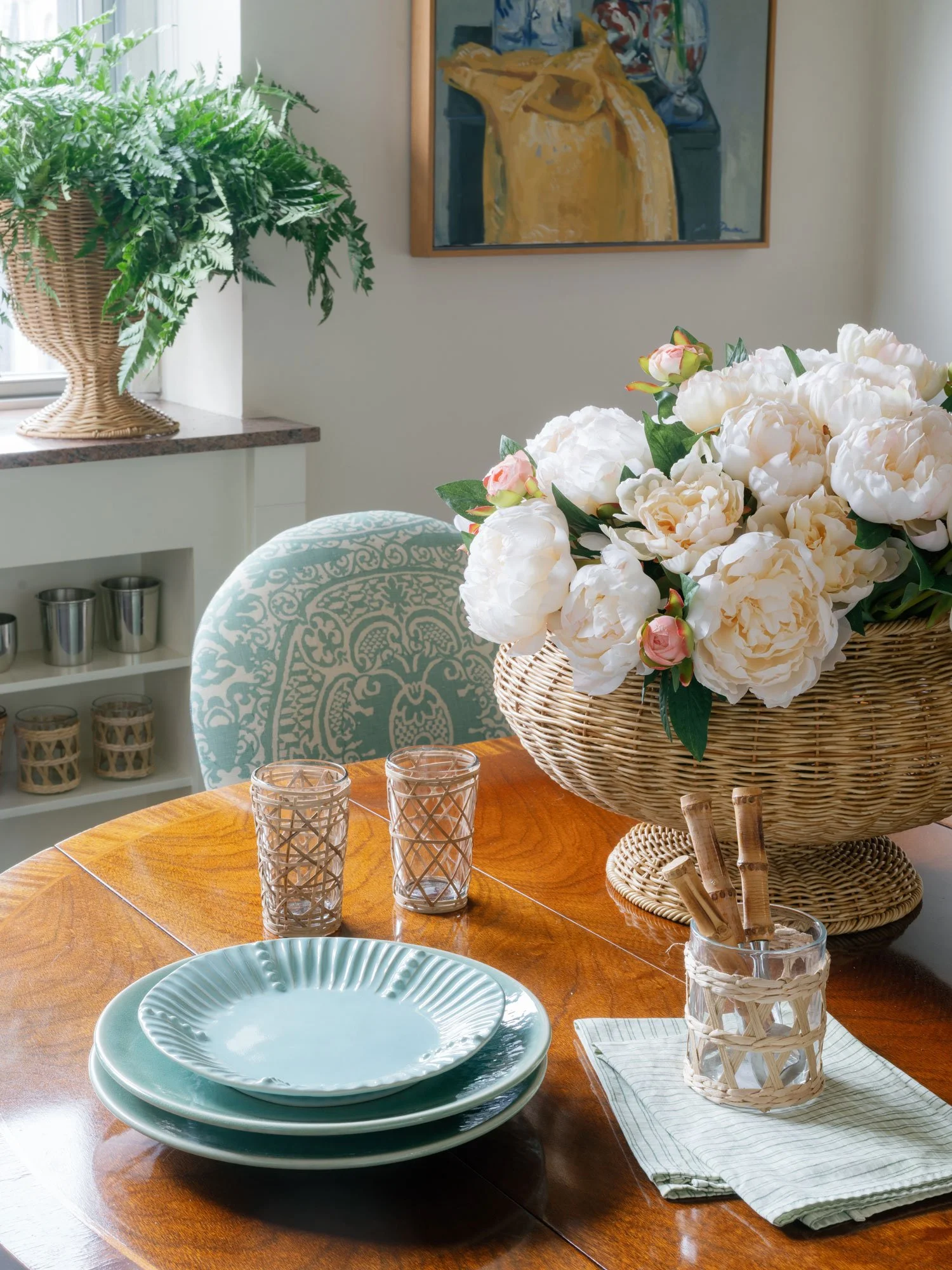 Close-up of dining table in New York City apartment by Elizabeth Hartz Interior Design featuring woven centerpiece basket with soft white and blush florals, aqua ceramic dinnerware, textured glass votives, and patterned upholstered dining chair in th