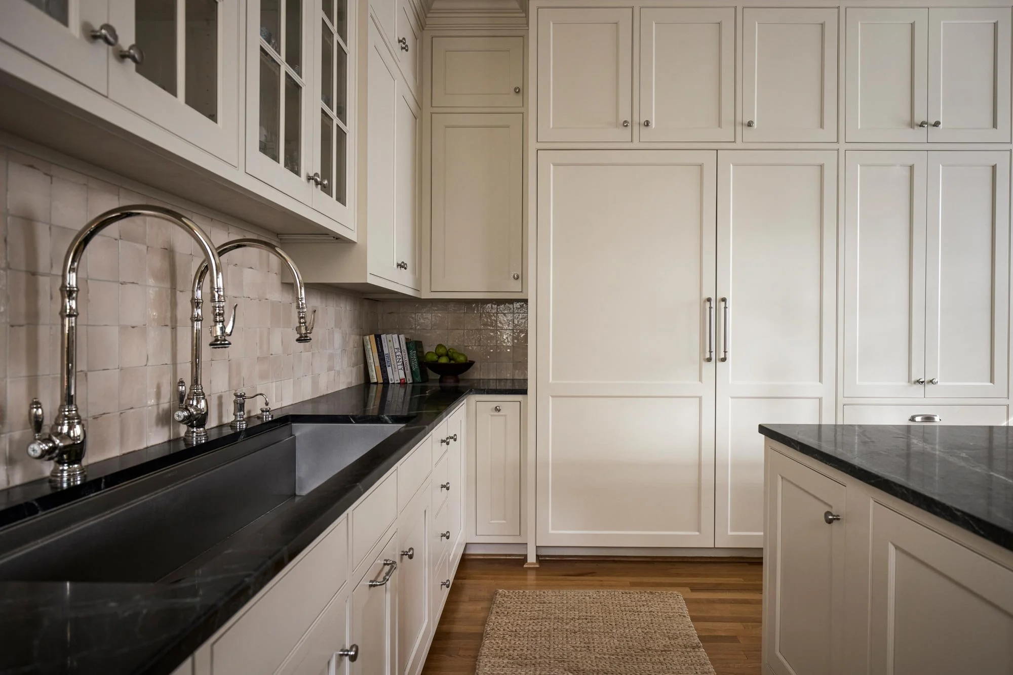 Kitchen with white cabinets, black countertops, a double sink with chrome faucets, a small black bowl with limes, some cookbooks, and a beige rug on wood flooring.
