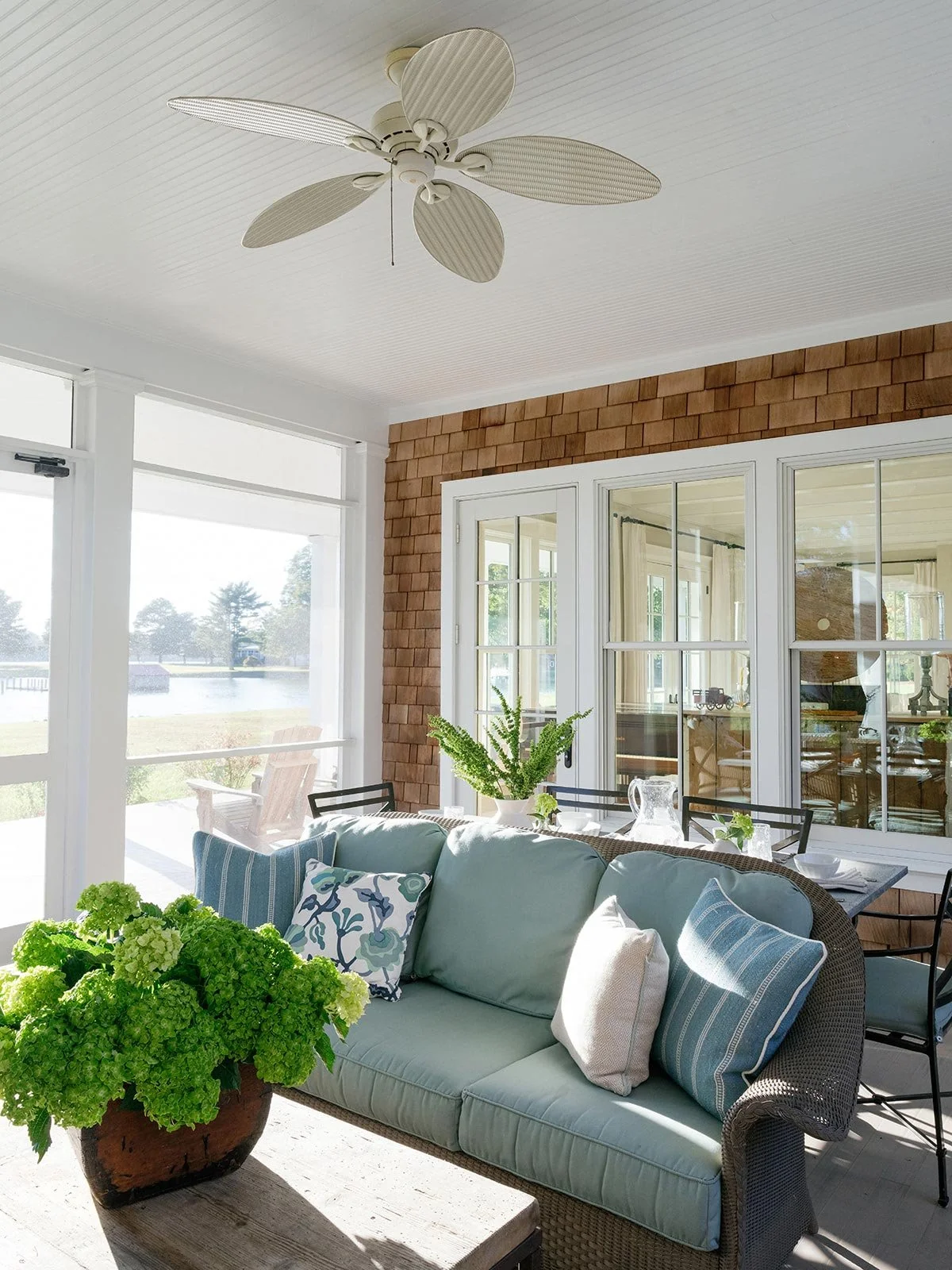 Enclosed porch designed by Elizabeth Hartz Interior Design at The River home in Kilmarnock, Virginia. Features wicker seating with blue cushions, coastal-patterned pillows, and greenery overlooking the water for a relaxed, elegant outdoor living spac