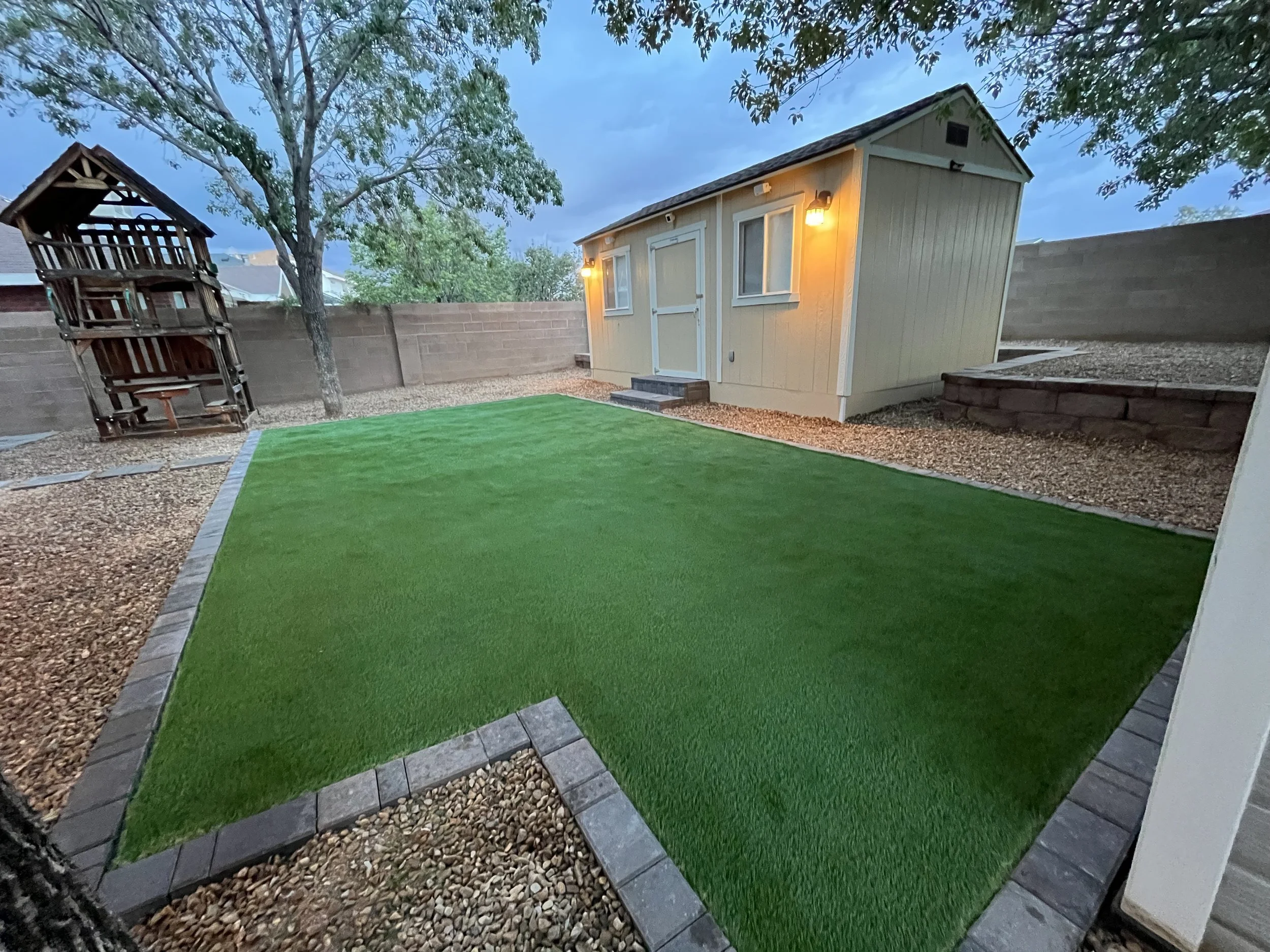 Backyard with a small shed, a tree, and a green artificial turf lawn, with outdoor lights on the shed and a wooden play structure.