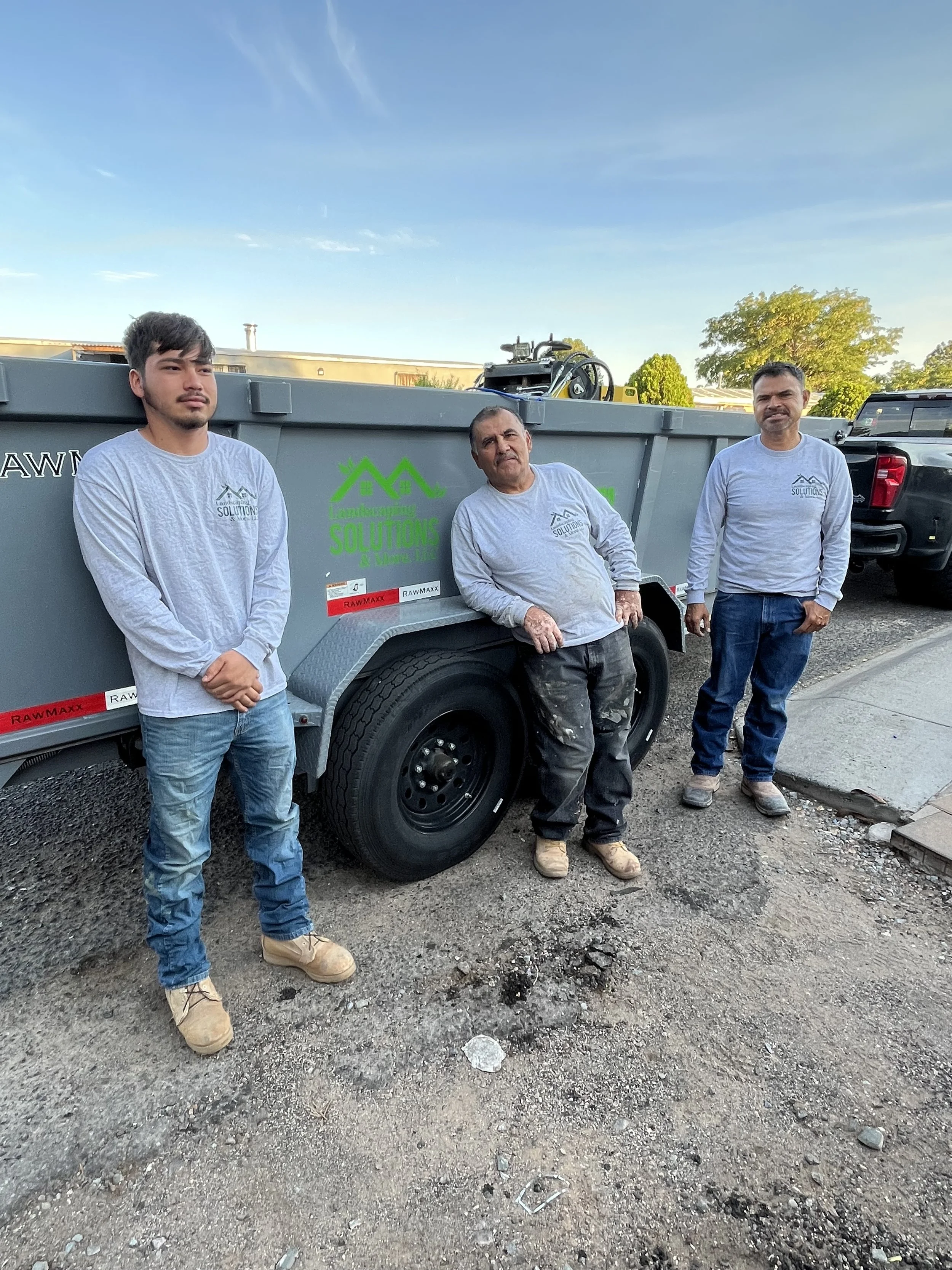 Three men in gray long sleeve shirts and jeans standing in front of a trailer with a green logo that reads "Larecycling Solutions". They are outdoors on a partly sunny day.