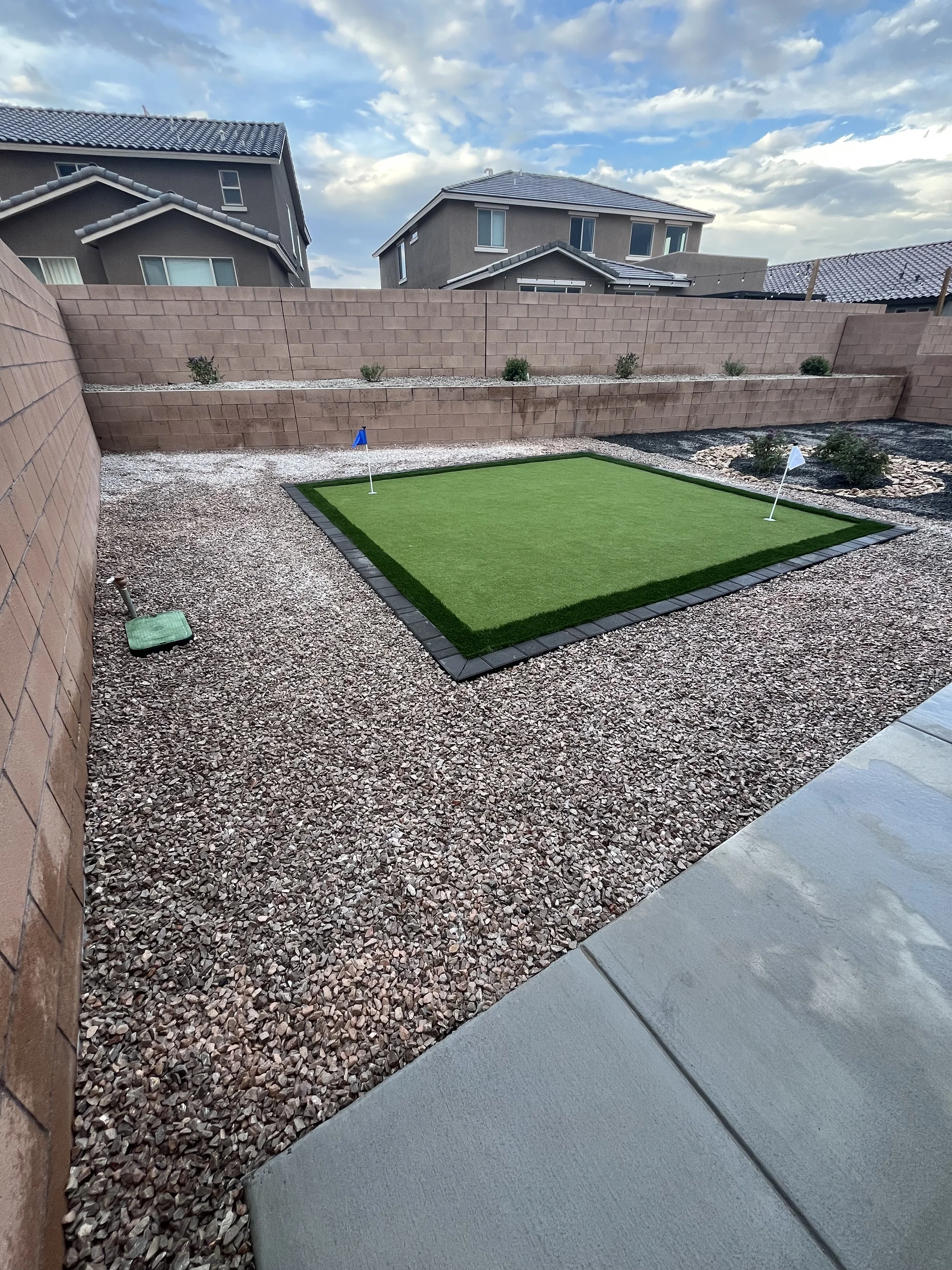 Backyard with small artificial putting green, gravel ground, brick wall fencing, and a few small plants against the wall. Houses and a partly cloudy sky are visible in the background.