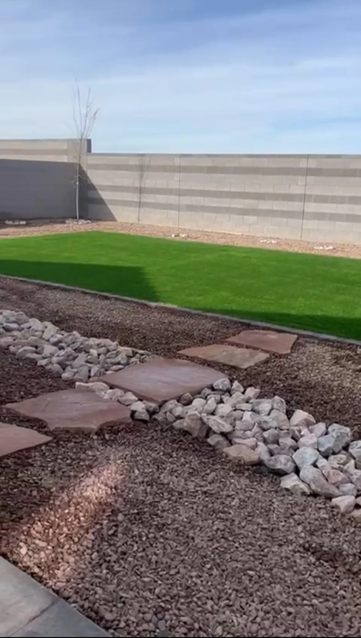 A landscaped backyard with a green grassy area, stepping stones, rocks, and a brick wall in the background.