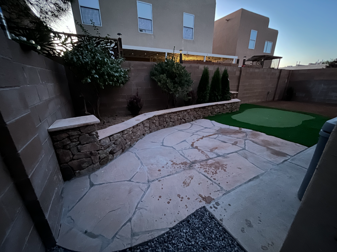 Backyard patio with stone and concrete flooring, a small artificial putting green, a raised garden bed with shrubs and trees, surrounded by a brick wall fence, with a modern house in the background during dusk.