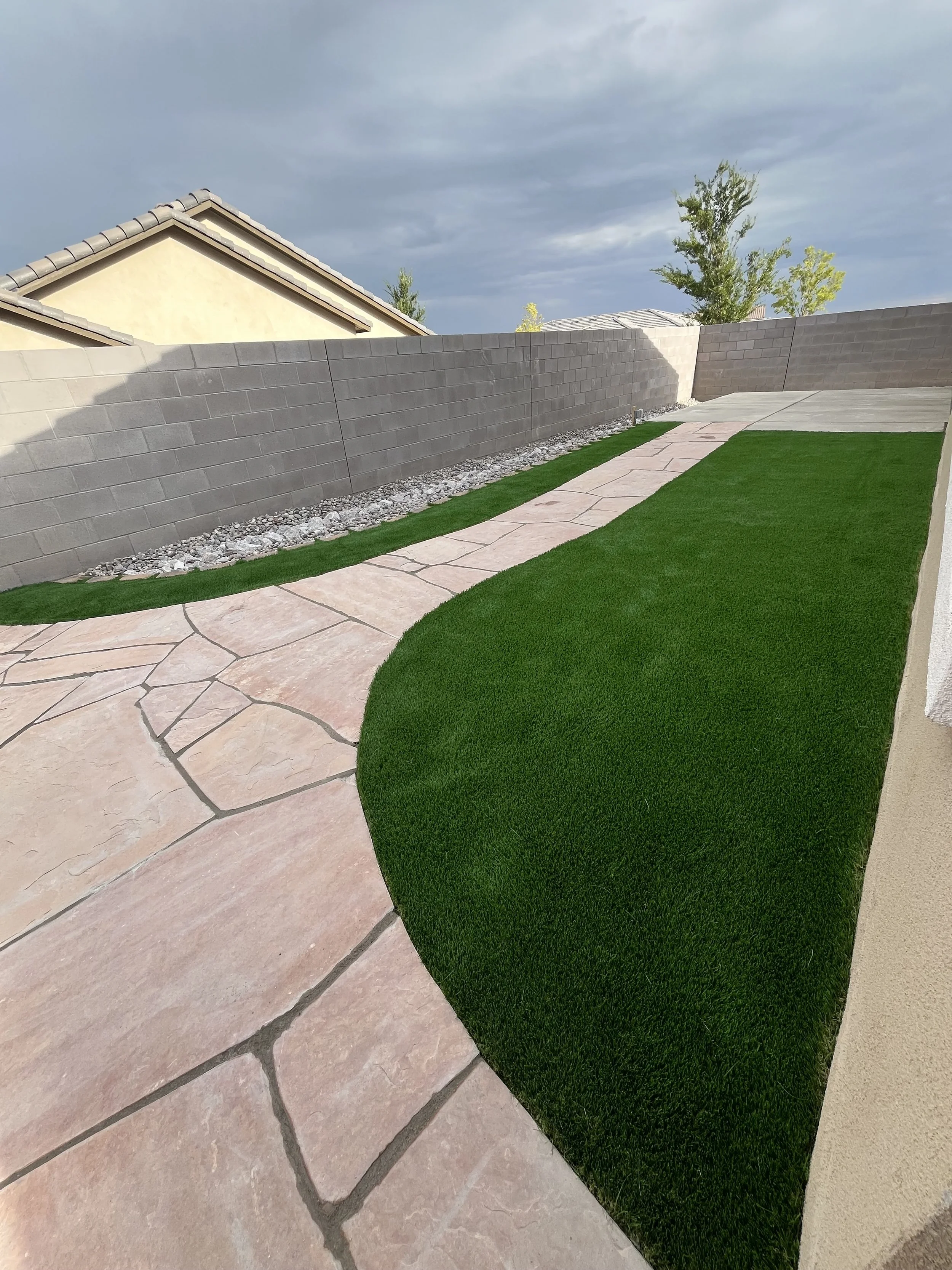 A backyard with a curved pinkish stone pathway, green grass, and a grey cinder block wall under a cloudy sky.