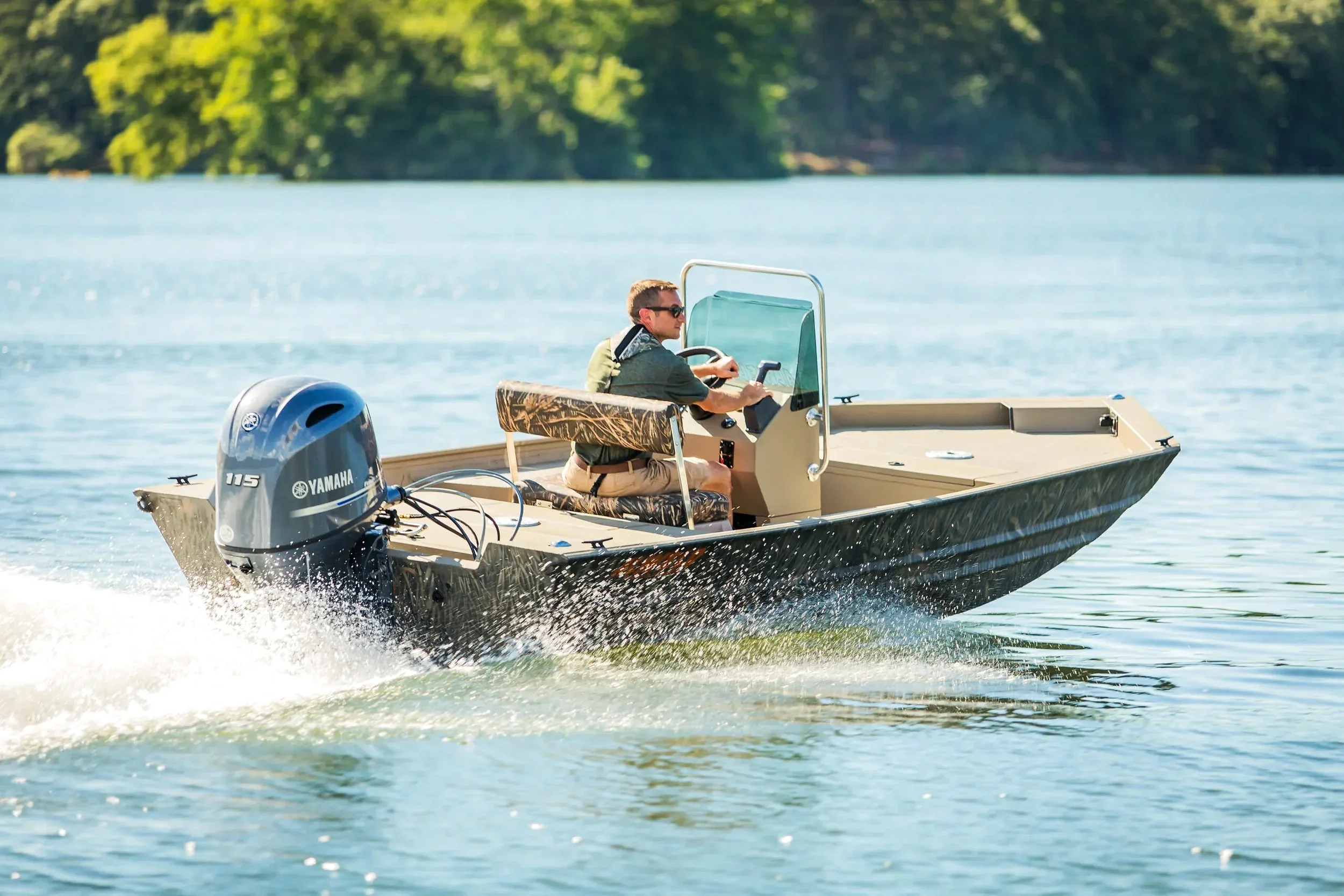 Man driving a camo-pattern aluminum alweld fishing boat with a Yamaha 115 outboard motor across a calm lake on a sunny day.