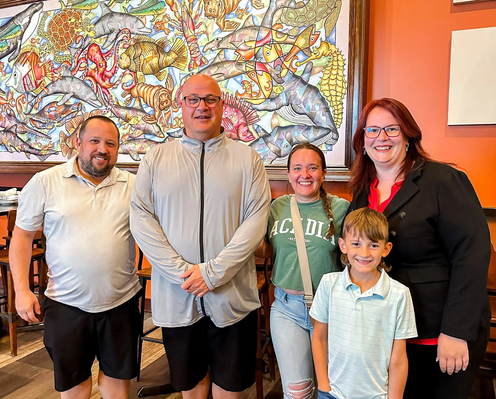 Group of five people smiling in a restaurant with a colorful fish-themed mural on the wall behind them.