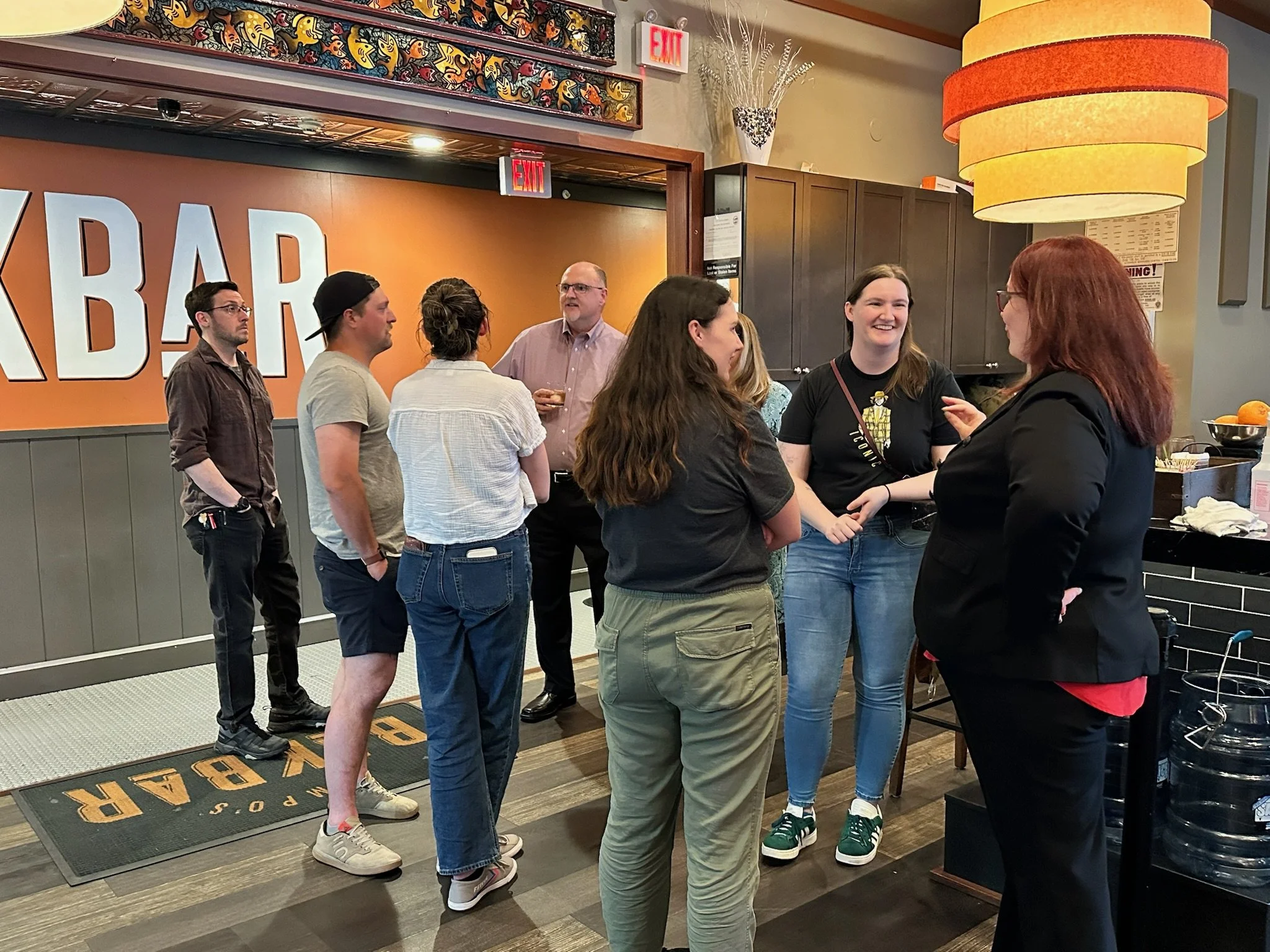 Group of people chatting in a bar with large orange and white sign reading 'BAKBAR'.