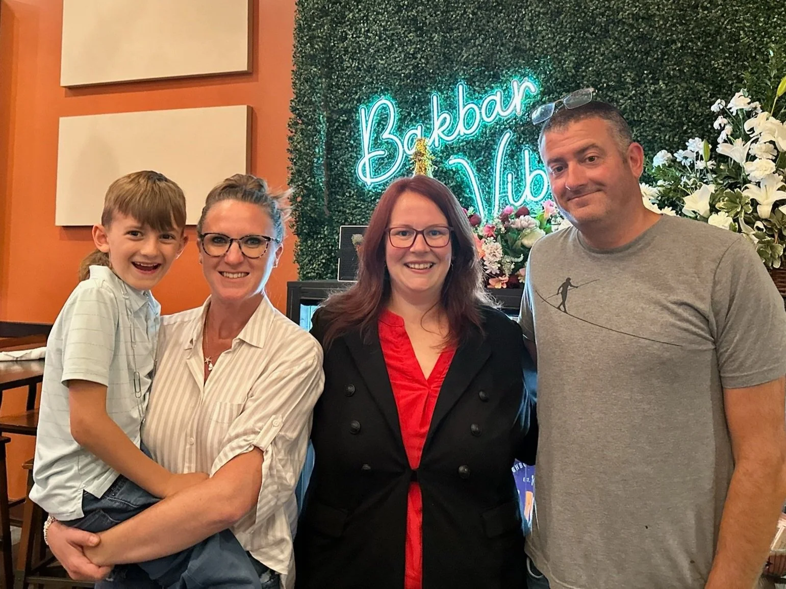 Four people smiling and posing for a photo in a restaurant with a neon sign that reads 'BakBar Vibes' and decorative flowers in the background.