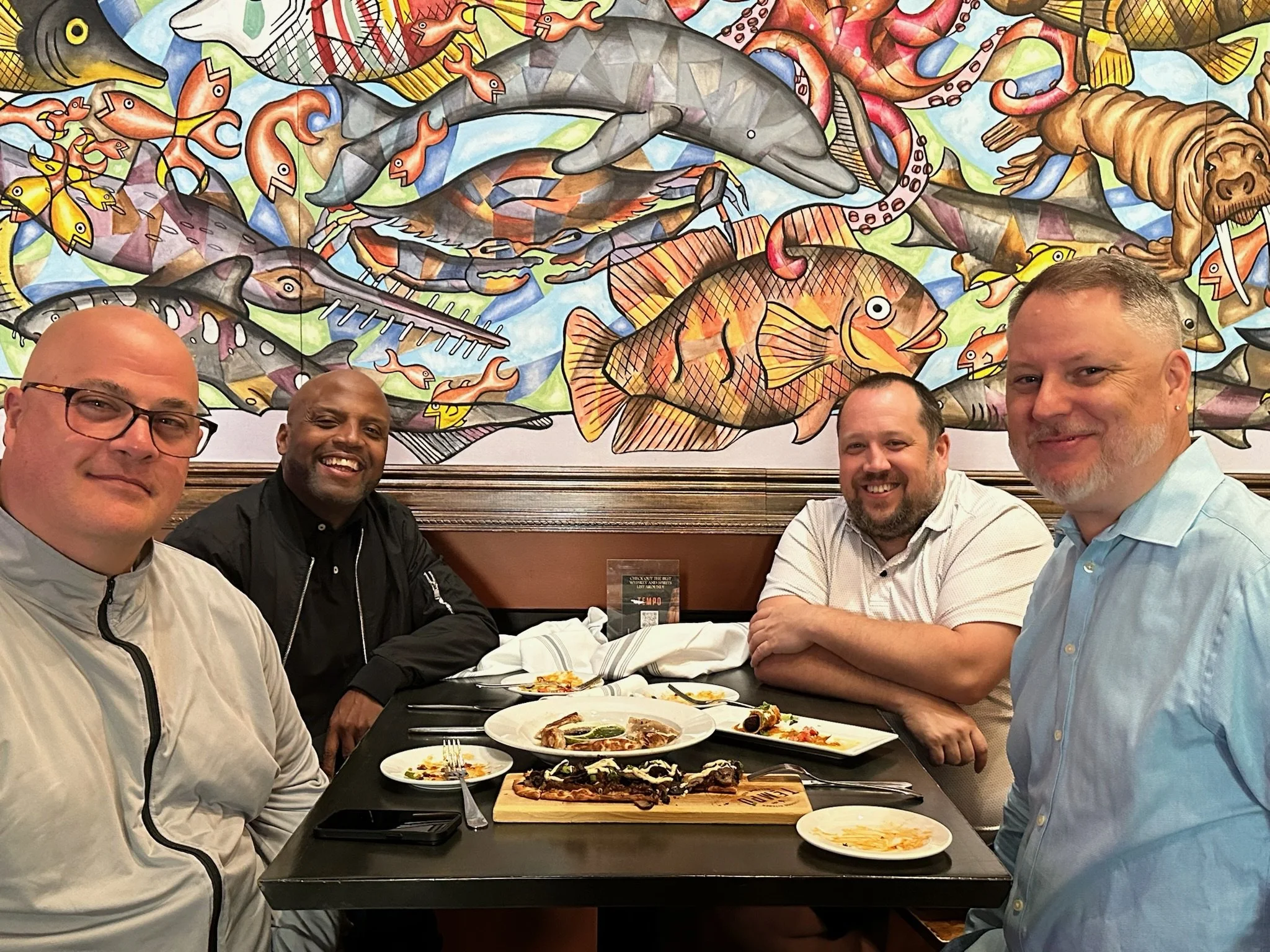 Four men sitting at a dining table inside a restaurant with a colorful mural of various fish on the wall behind them. The table has plates of food, and all four men are smiling at the camera.